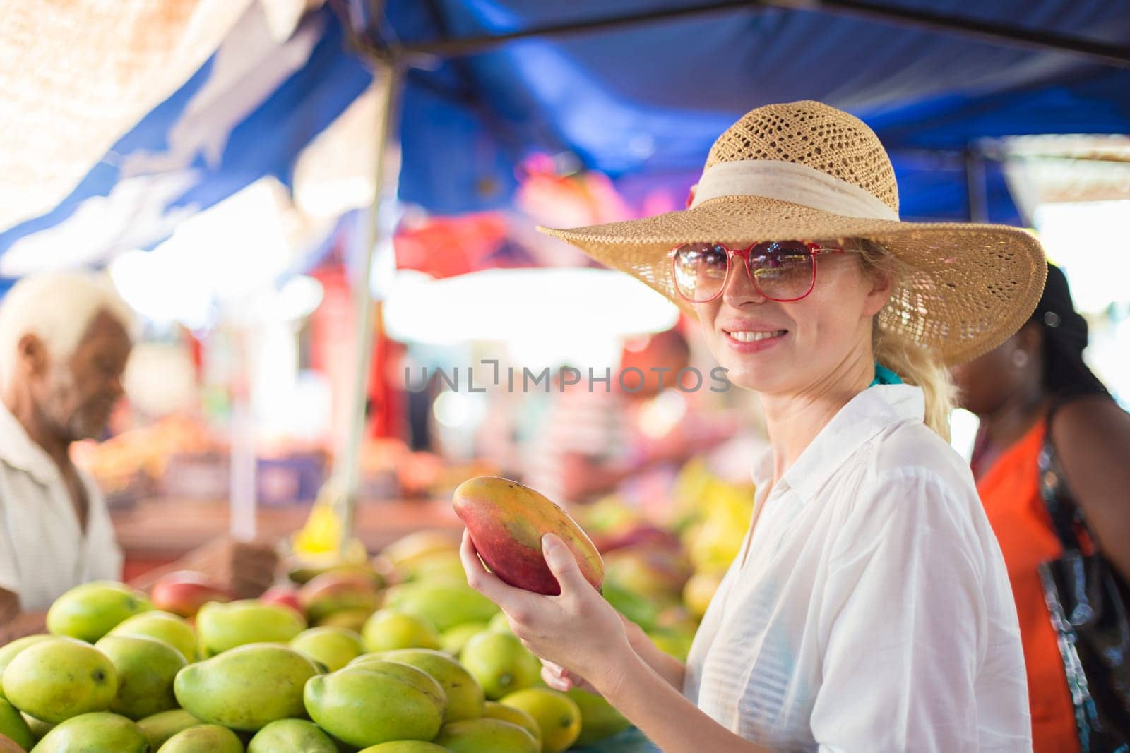 Female traveler wearing elegant colonial style white tunic and hat buying fresh tropical fruit on traditional Victoria food market on Seychelles islands.