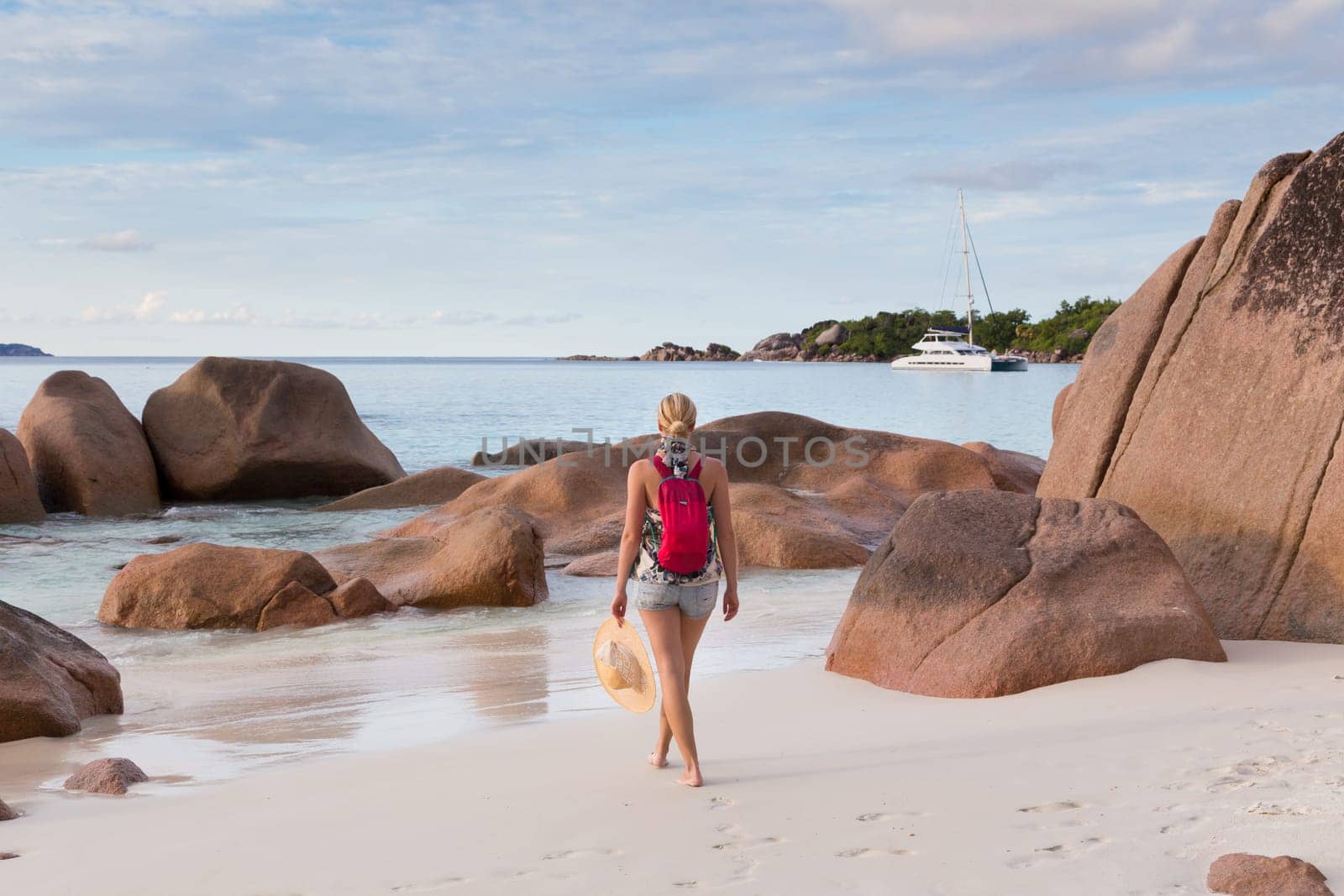 Woman wearing smal backpack, scarf, jeans shorts and beach hat, enjoying view of Anse Lazio beach on Praslin Island, Seychelles. Summer vacations on picture perfect tropical beach concept.