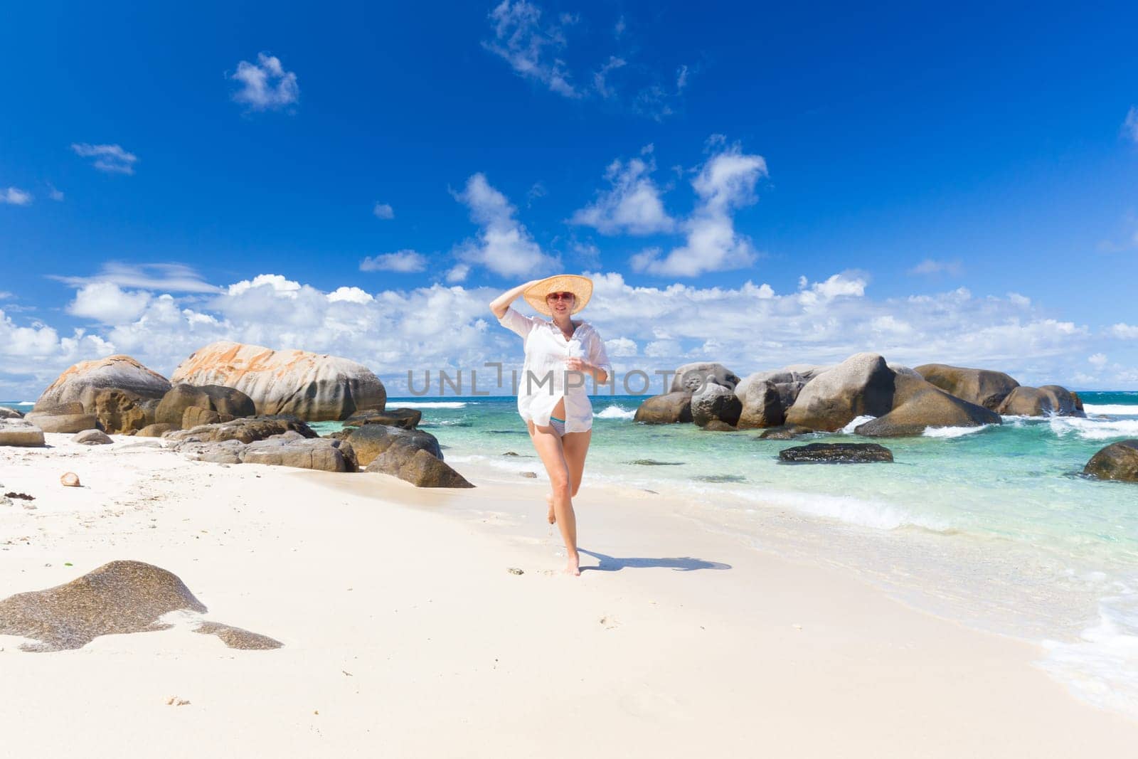 Happy woman wearing white loose tunic over bikini and beach hat, enjoying amazing white sandy beach on Mahe Island, Seychelles. Summer vacations on picture perfect tropical beach concept.