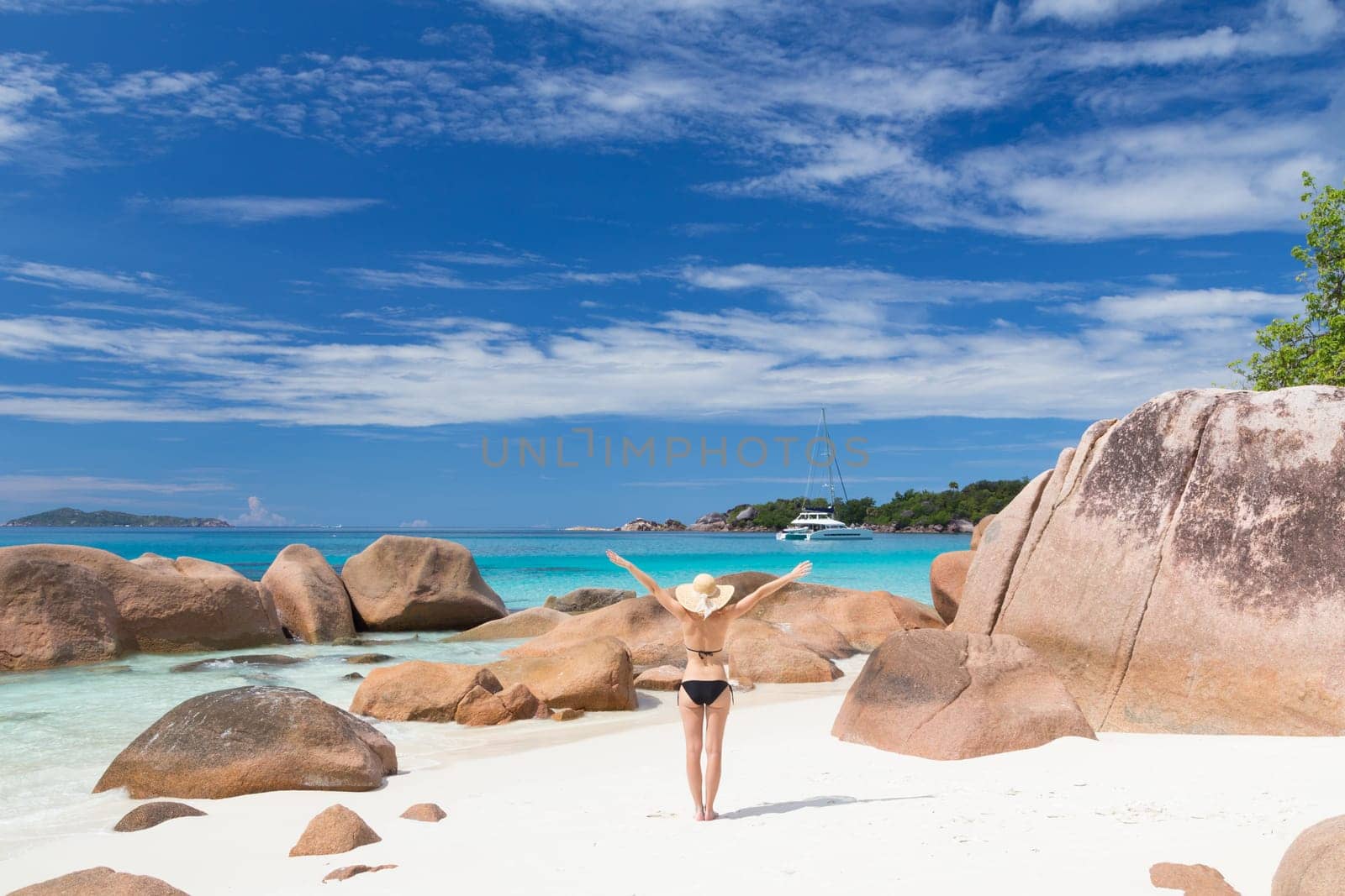 Woman arms rised, wearing black bikini and beach hat, enjoying amazing view on Anse Lazio beach on Praslin Island, Seychelles. Summer vacations on picture perfect tropical beach concept.