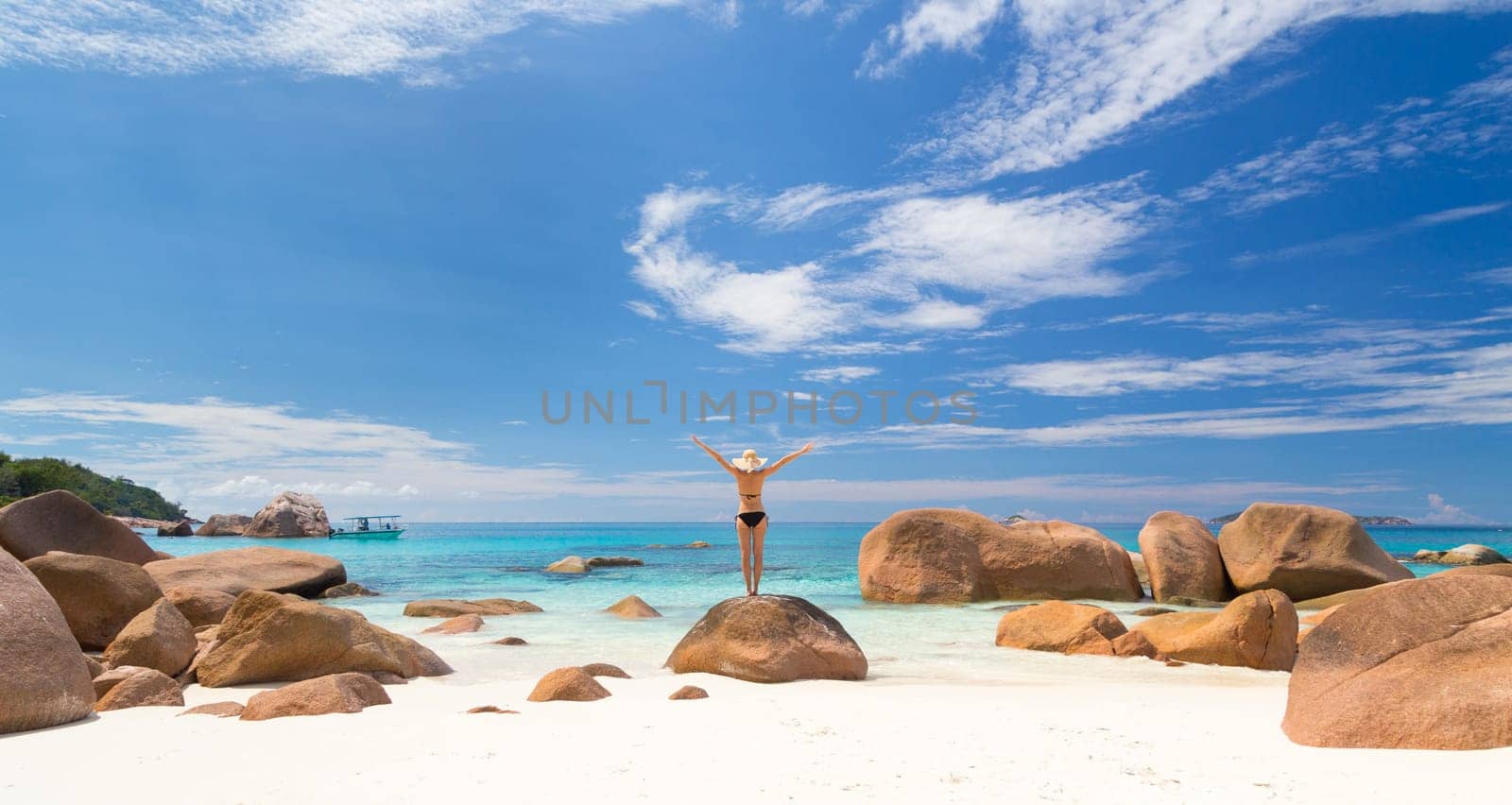 Woman, arms rised, wearing black bikini and beach hat, enjoying amazing view on Anse Lazio beach on Praslin Island, Seychelles. Summer vacations on picture perfect tropical beach concept. Copy space.