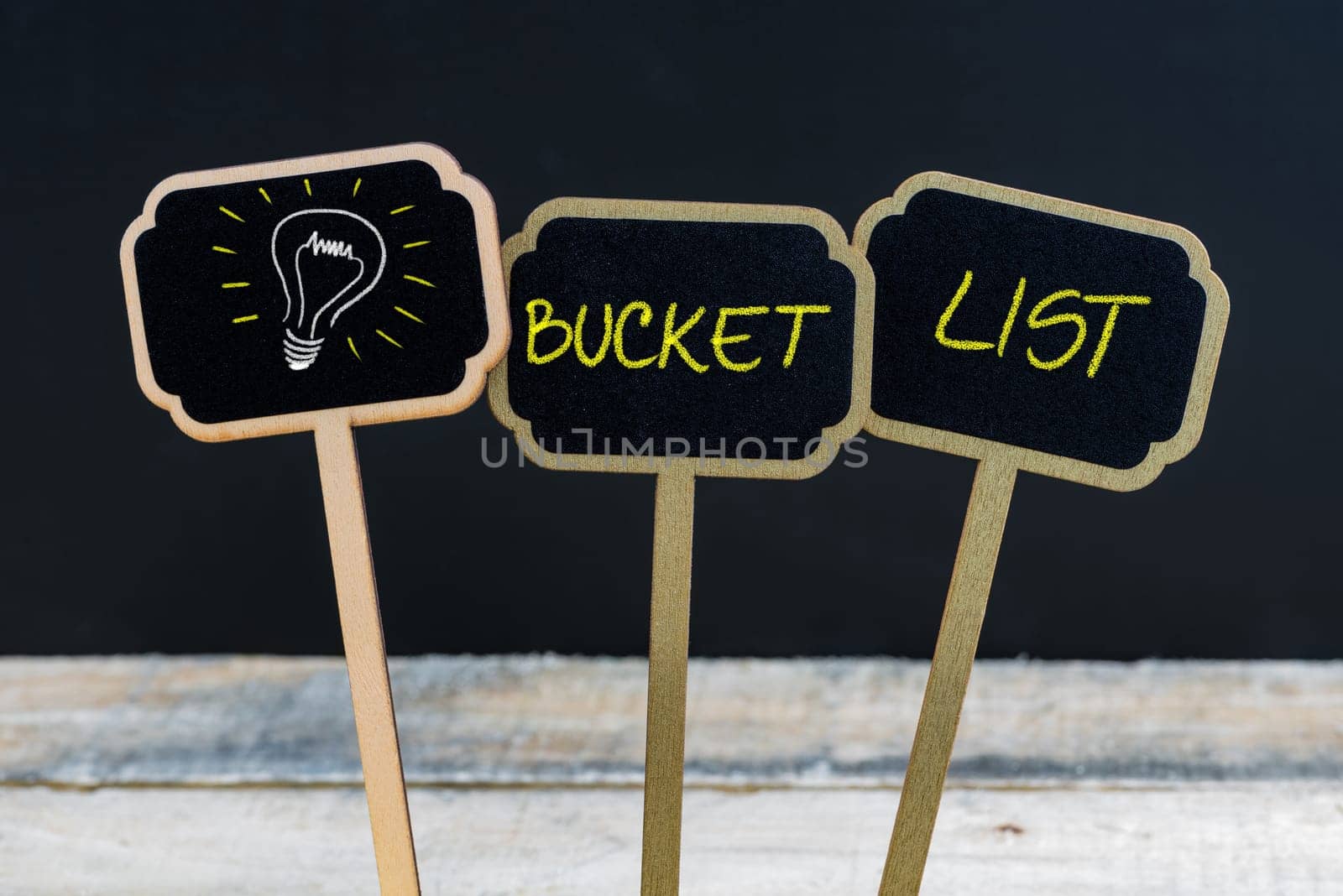Concept message BUCKET LIST and light bulb as symbol for idea written with chalk on wooden mini blackboard labels, defocused chalkboard and wood table in background