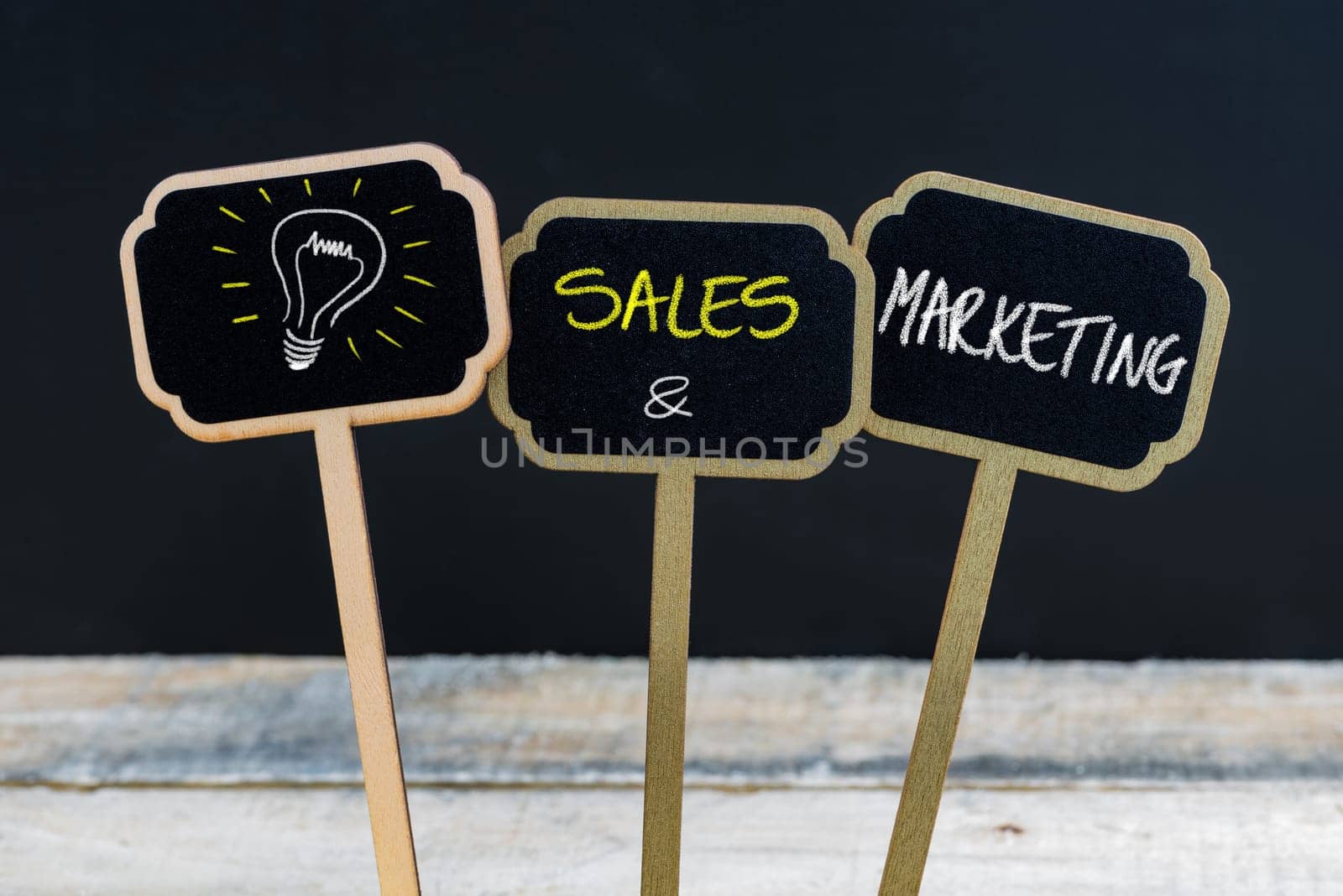 Concept message SALES AND MARKETING and light bulb as symbol for idea written with chalk on wooden mini blackboard labels, defocused chalkboard and wood table in background