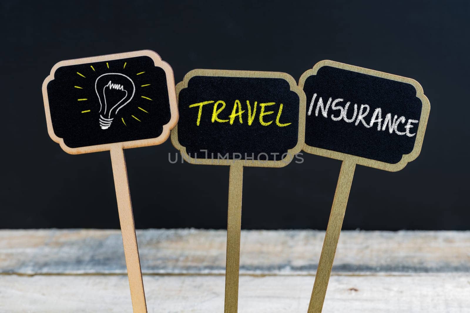 Concept message TRAVEL INSURANCE and light bulb as symbol for idea written with chalk on wooden mini blackboard labels, defocused chalkboard and wood table in background