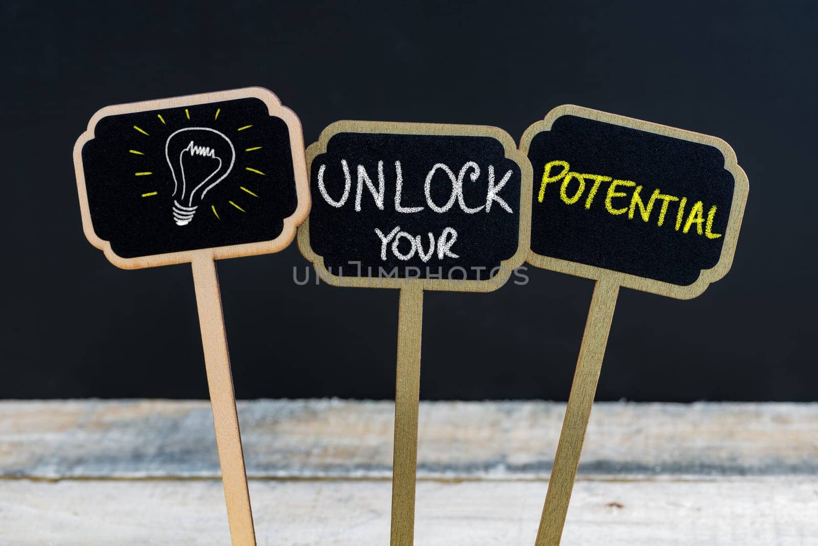 Concept message UNLOCK YOUR POTENTIAL and light bulb as symbol for idea written with chalk on wooden mini blackboard labels, defocused chalkboard and wood table in background