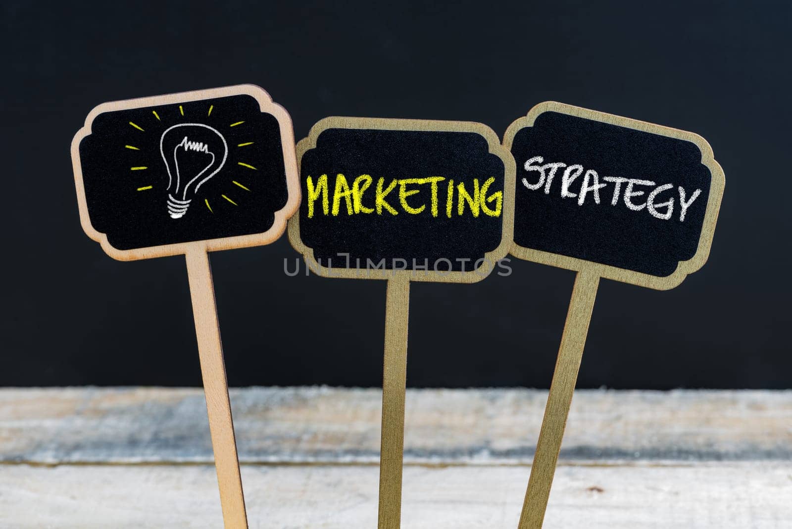 Concept message MARKETING STRATEGY and light bulb as symbol for idea written with chalk on wooden mini blackboard labels, defocused chalkboard and wood table in background