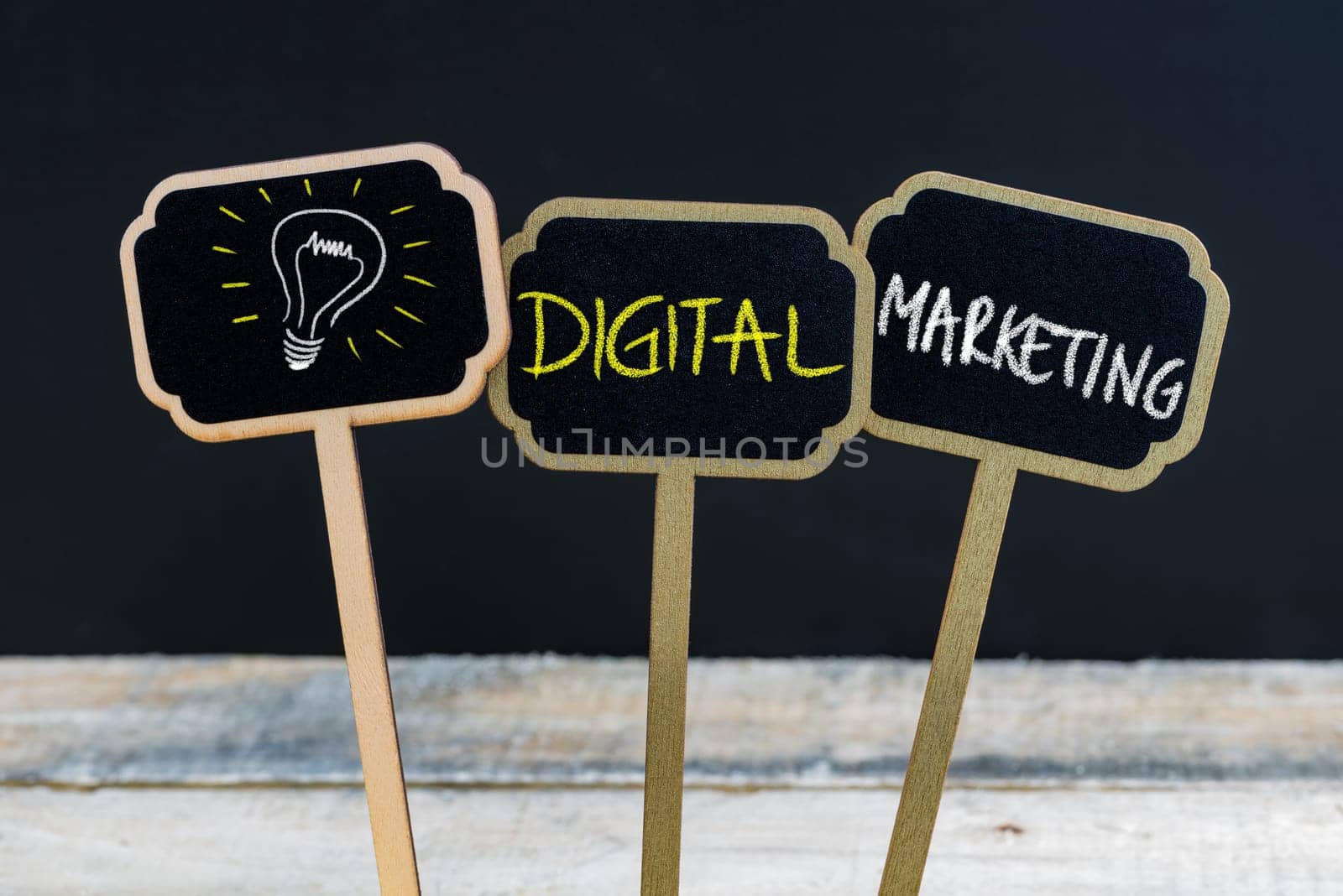 Concept message DIGITAL MARKETING and light bulb as symbol for idea written with chalk on wooden mini blackboard labels, defocused chalkboard and wood table in background