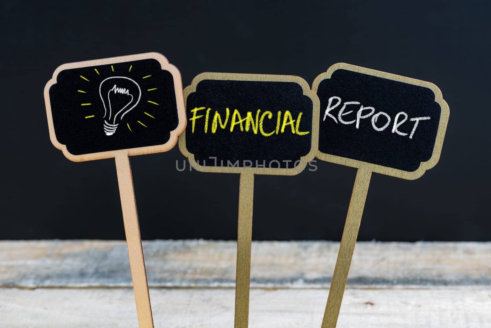 Concept message FINANCIAL REPORT and light bulb as symbol for idea written with chalk on wooden mini blackboard labels, defocused chalkboard and wood table in background