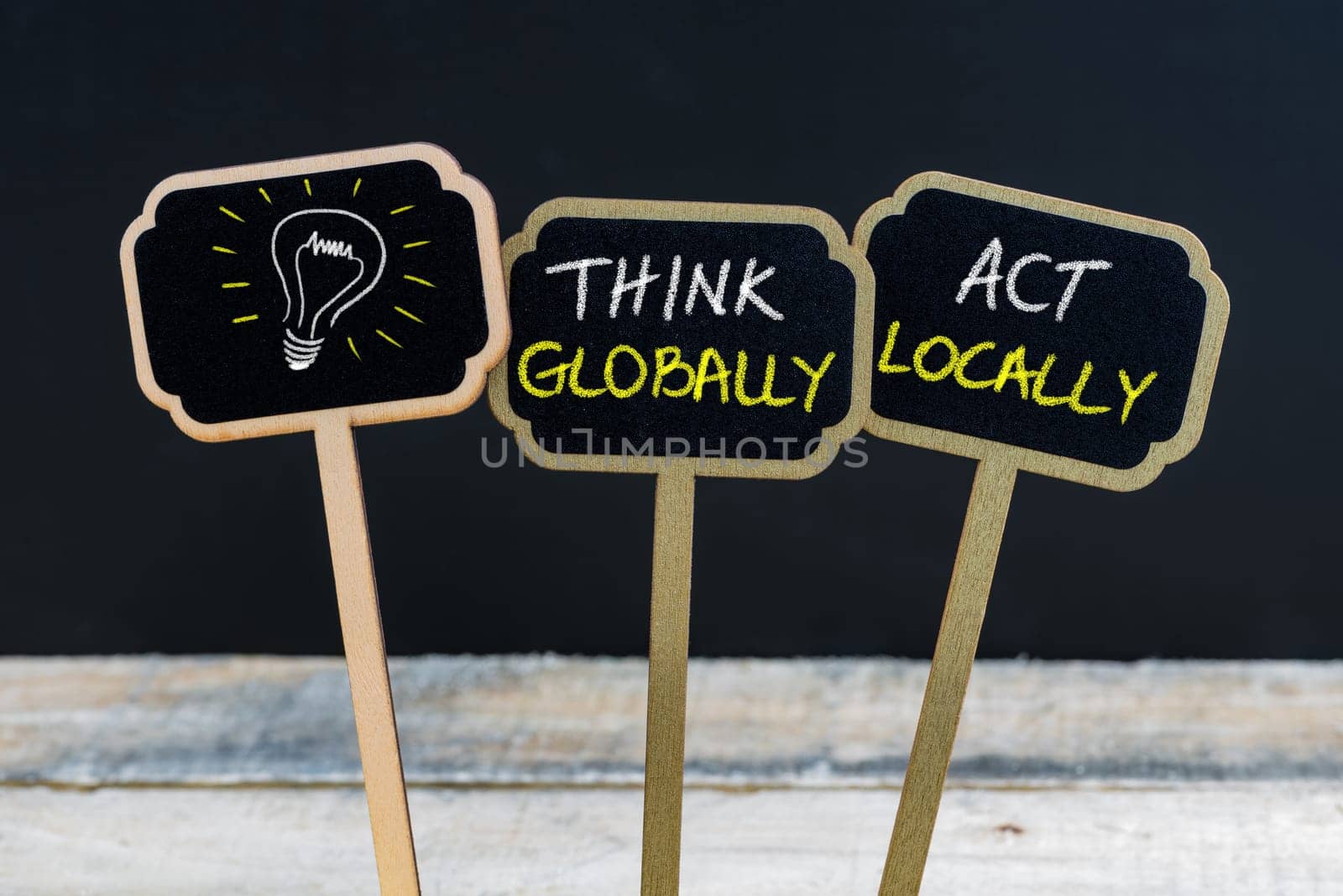 Concept message THINK GLOBALLY ACT LOCALLY and light bulb as symbol for idea written with chalk on wooden mini blackboard labels, defocused chalkboard and wood table in background