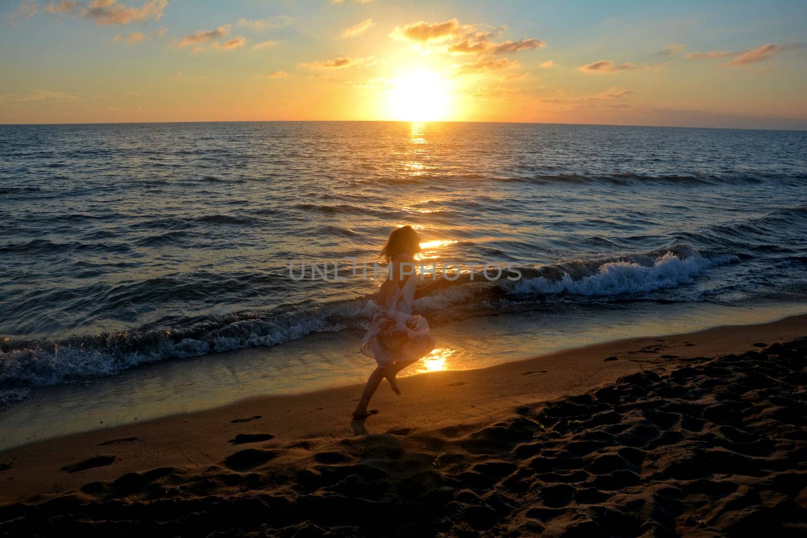Young girl in long flowers - dress runs barefoot at sunset along the beach