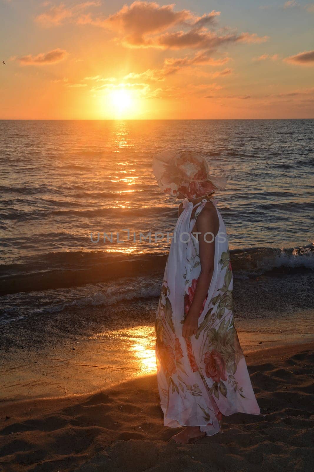 Girl in a long dress and hat on her head stands on the sea beach at sunset,with a view of the sun