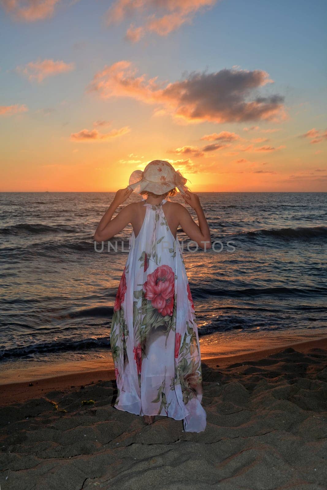 Girl in a long dress and holds hat on her head with both hands and stands on the sea beach at sunset,with a view to the sun,head covers the sun