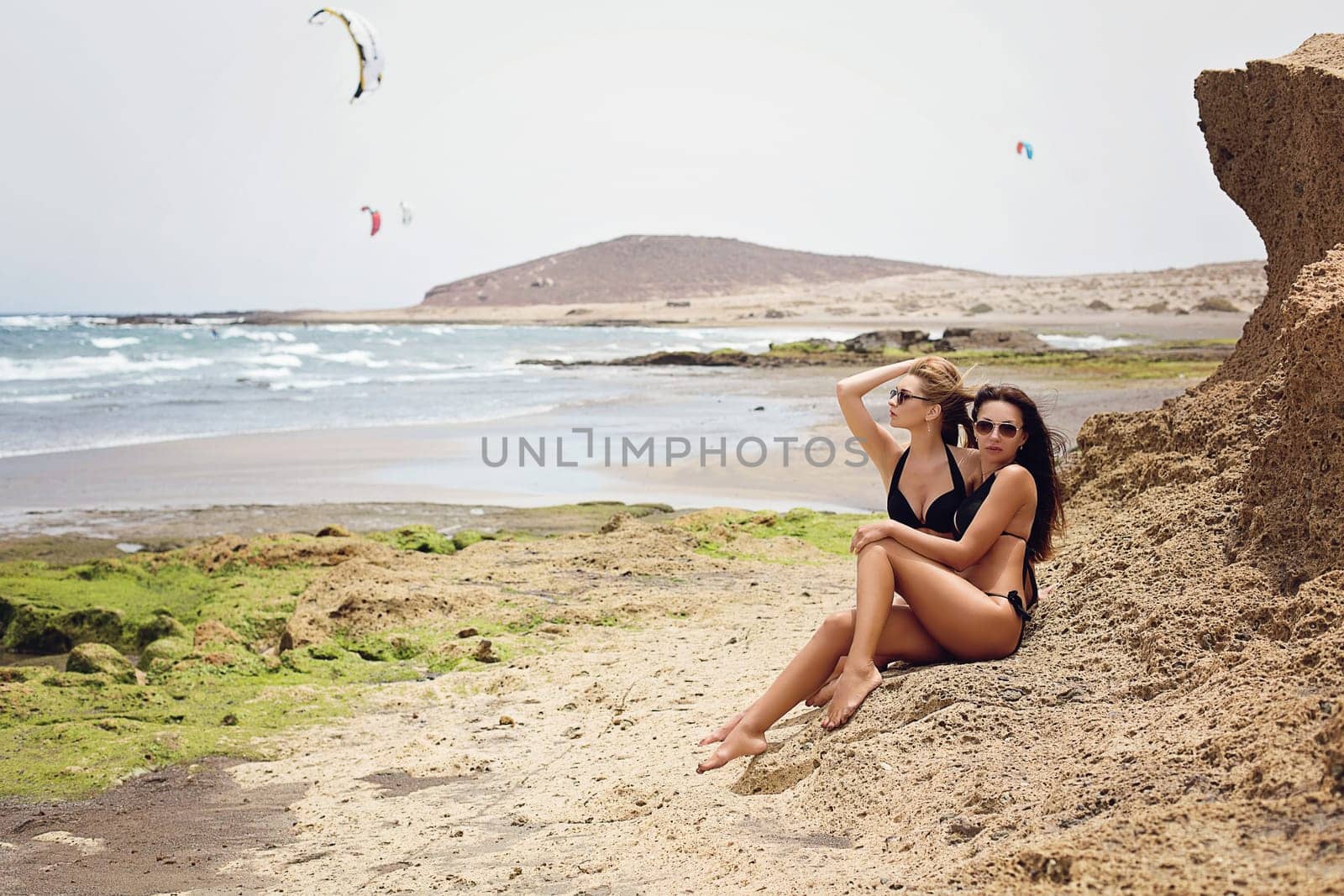 Two beautiful women sitting on wild ocean beach and watching parasurfers. Tenerife. Outdoor shot.