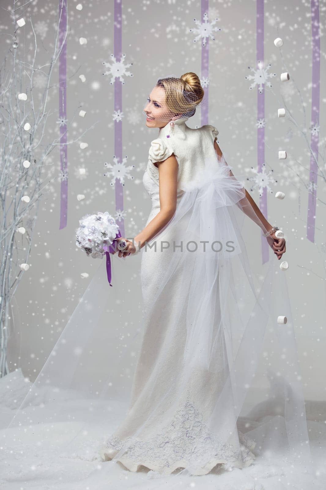 Portrait of beautiful young woman in long warm gown for winter wedding with purple flower bouquet. Over snowy background