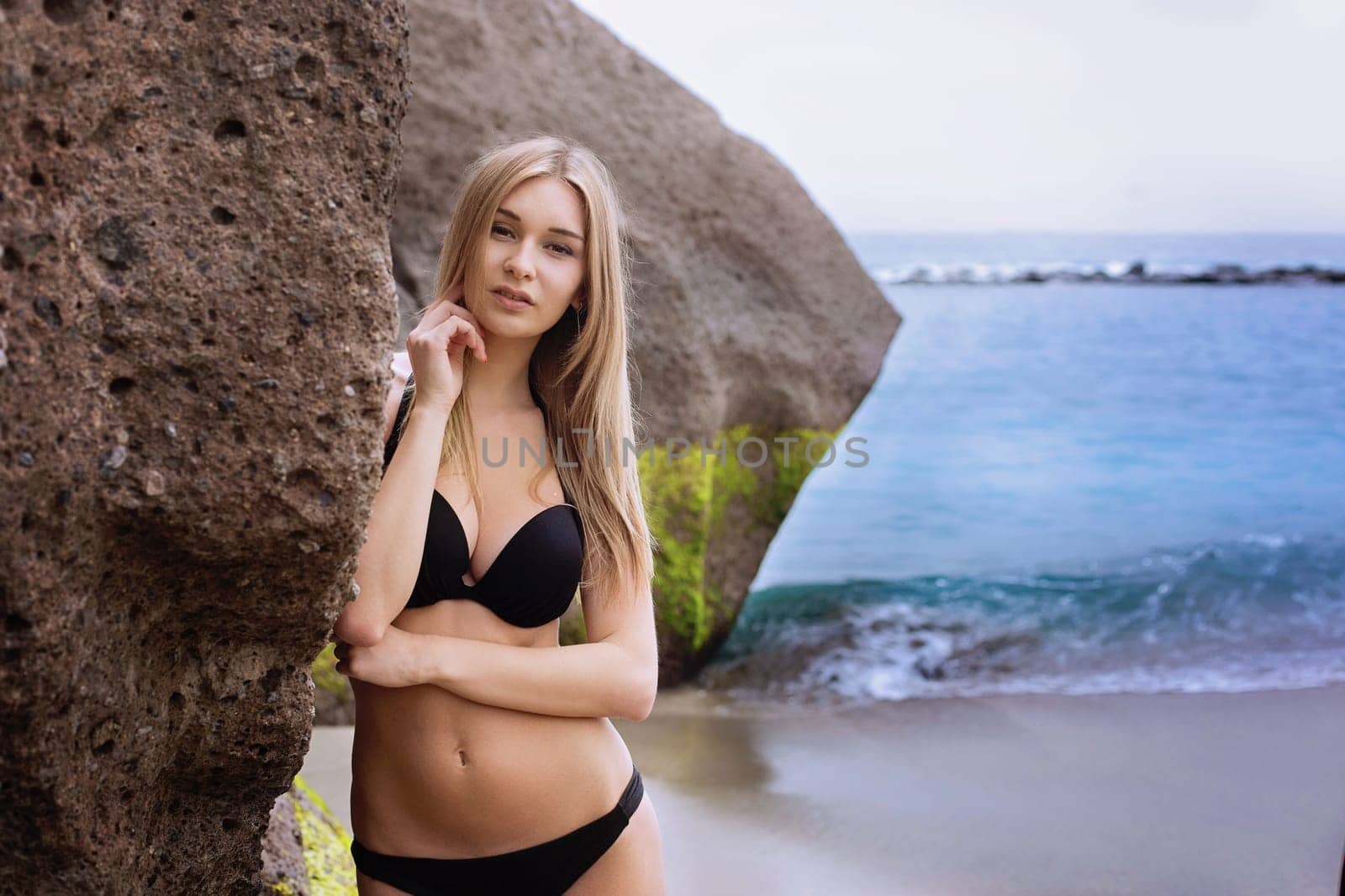 Beautiful young woman with long blond hait in black bikini standing near cliff. Outdoor shot. Tenerife.