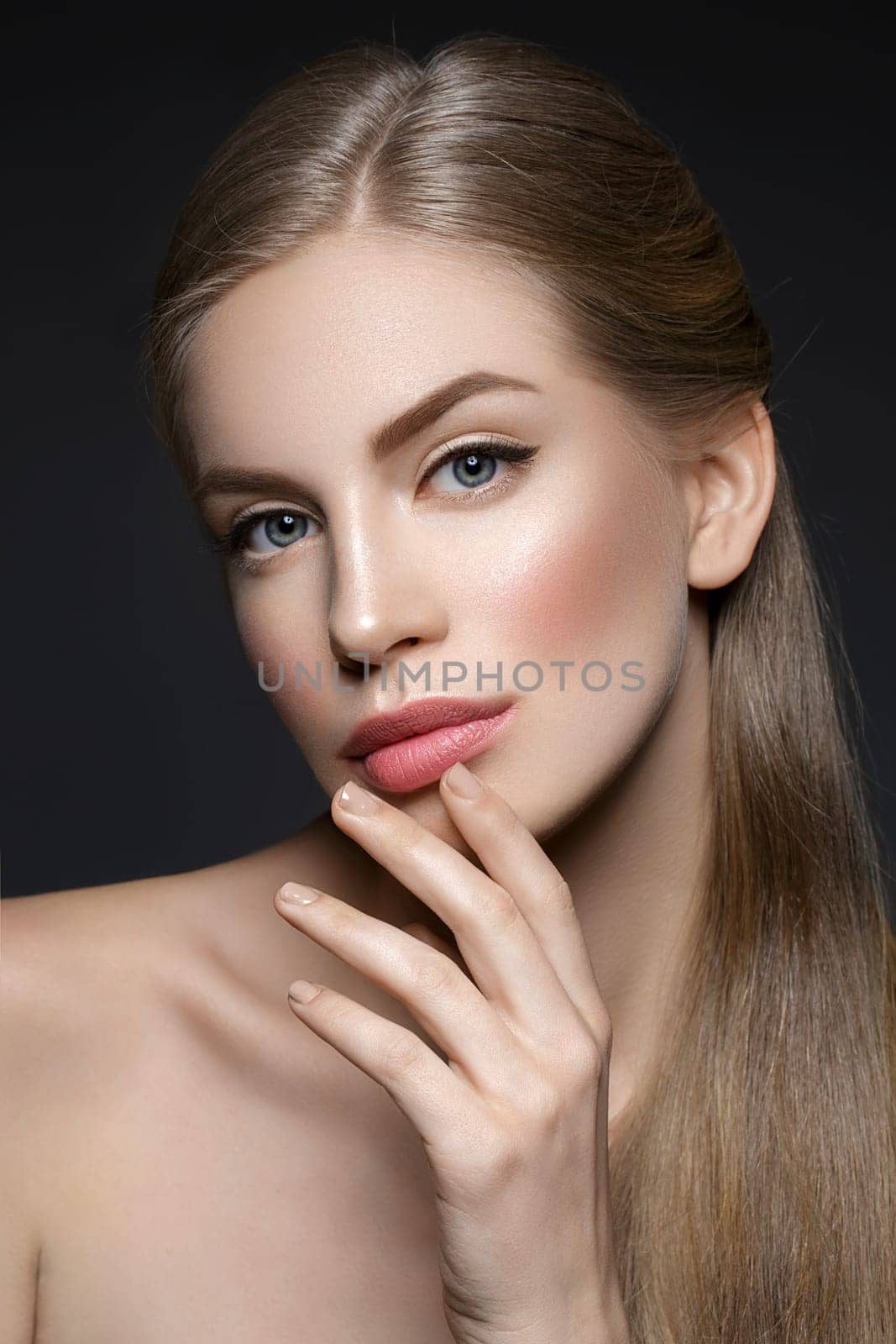 Beautiful young woman with blond hair and nude pink lips touching face. Over black background. Closeup beauty shot.