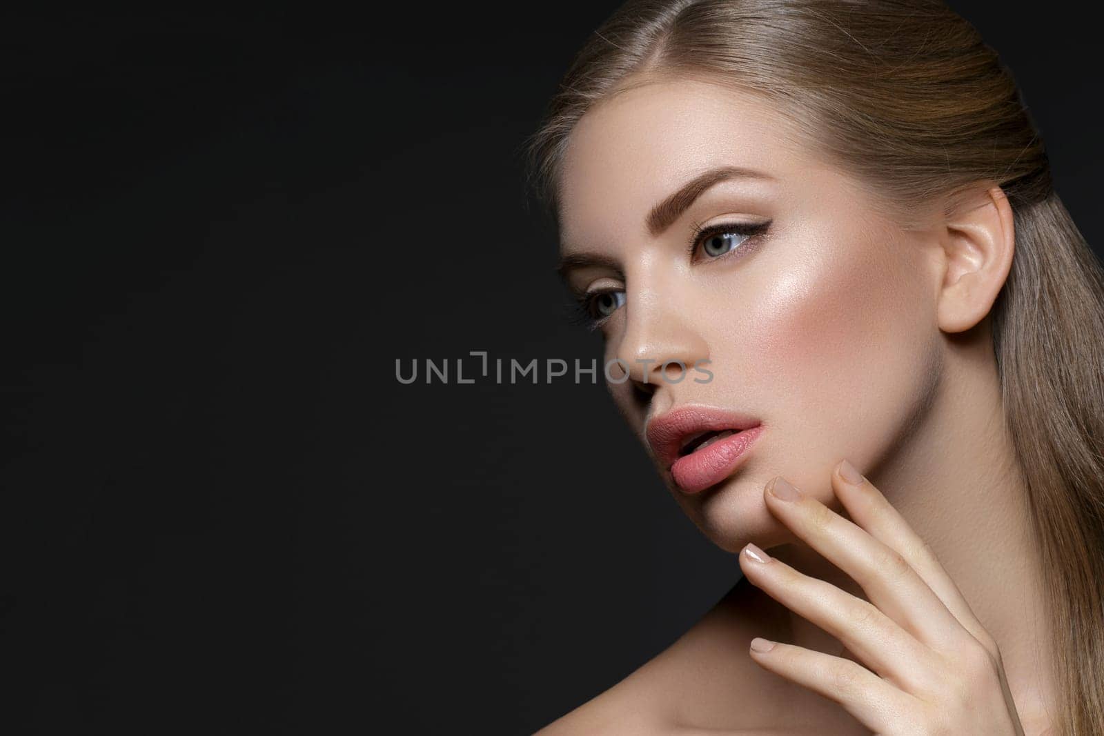Beautiful young woman with blond hair and nude pink lips touching face. Over black background. Closeup beauty shot.