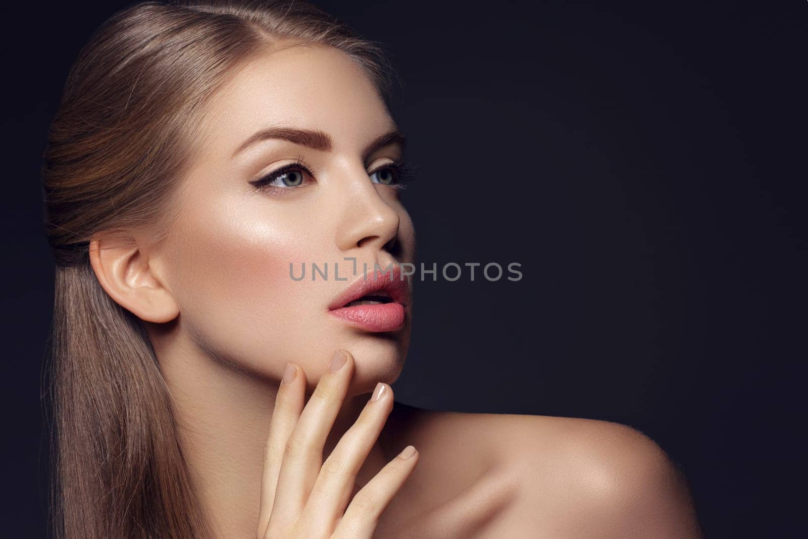 Beautiful young woman with blond hair and nude pink lips touching face. Over black background. Closeup beauty shot.