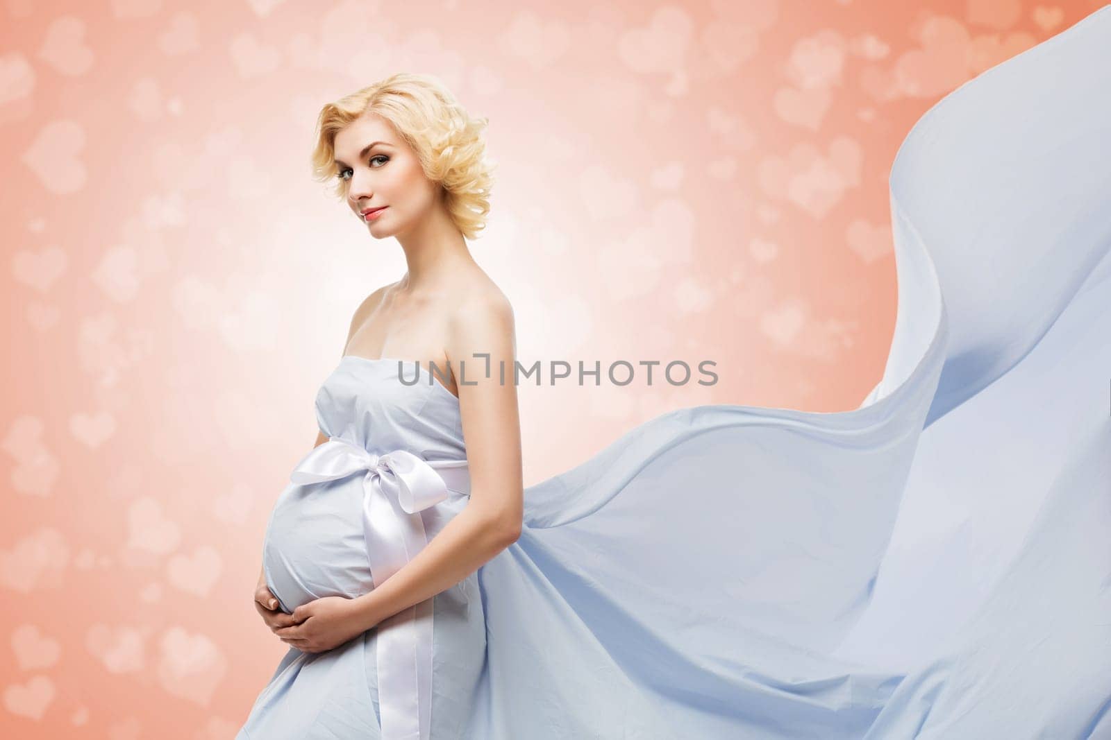 Beautiful young pregnant woman in blue flying fabrics standing over coral background with heart bokeh on it. Isolated.