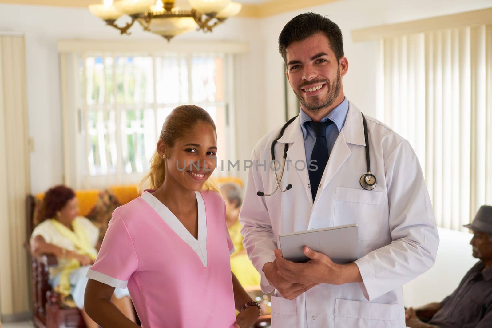 Portrait of doctor and nurse at work in hospital for seniors. The man holds a tablet pc. Some elderly people are sitting in background