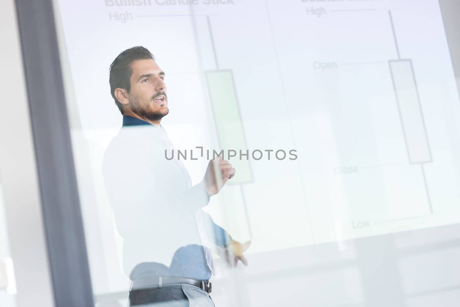 Business man making a presentation in front of whiteboard. Business executive delivering a presentation to his colleagues during meeting or in-house business training. View through glass.