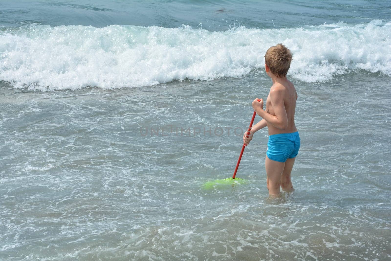 Little blond boy with blue swimming trunks stands in the water with a net and waits for the sea wave
