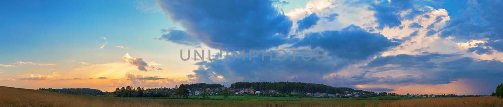 Sunset in the countryside. Dramatic sky with clouds, field and rural houses on a background of trees. Panoramic shot.
