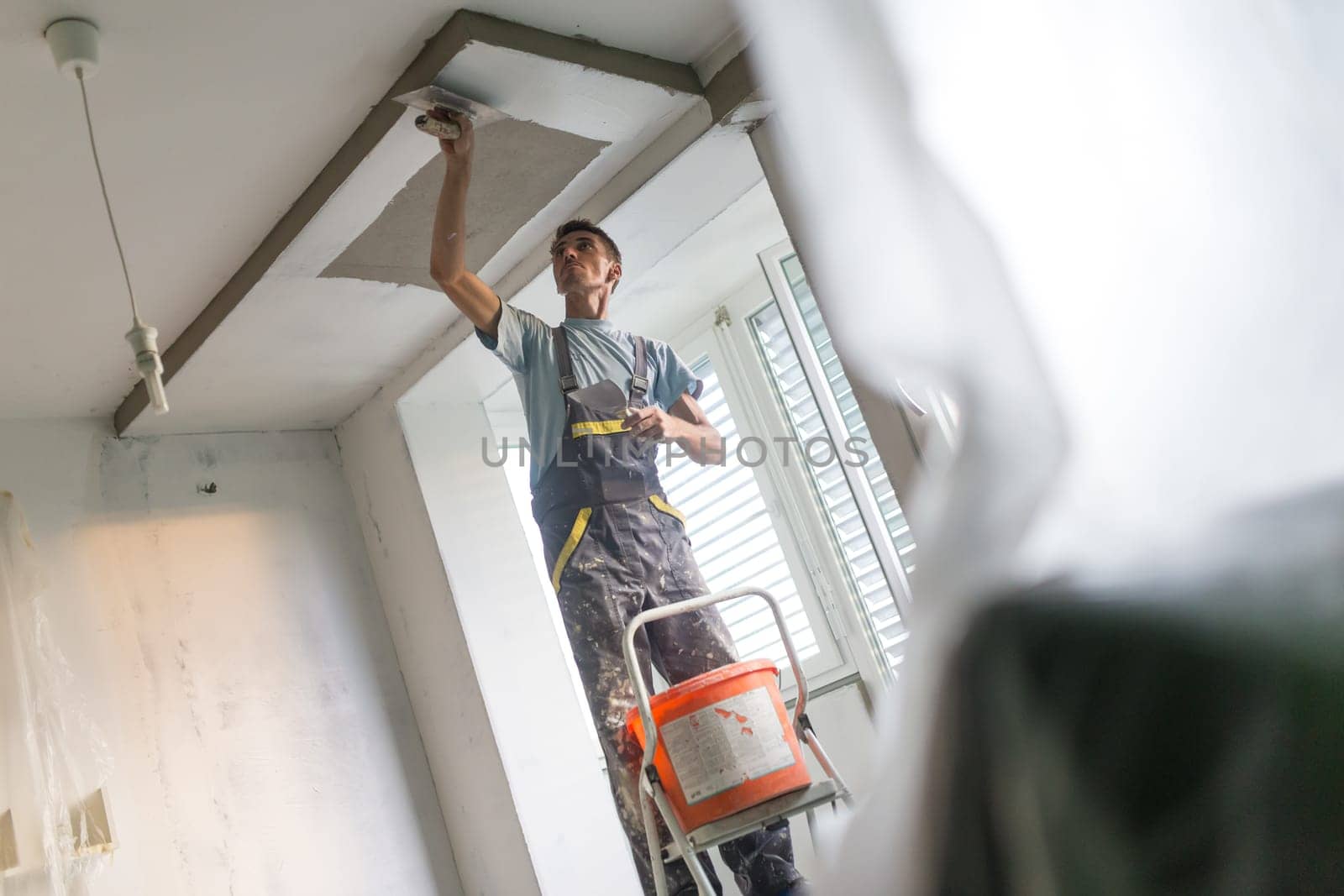 Construction worker wearing worker overall with wall plastering tools renovating apartment house. Plasterer renovating indoor walls and ceilings with float and plaster. Construction finishing works.