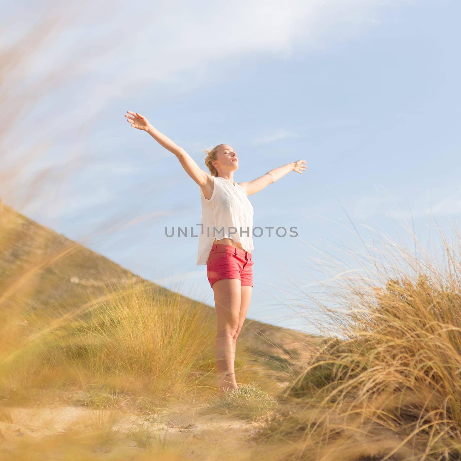 Relaxed woman, arms rised, enjoying sun, freedom and life an a beautiful beach. Young lady feeling free, relaxed and happy. Concept of vacations, freedom, happiness, enjoyment and well being.