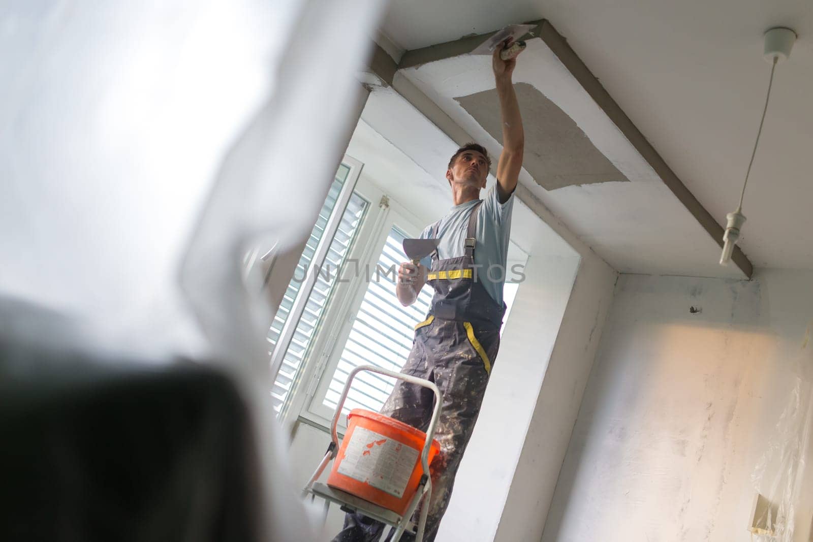 Construction worker wearing worker overall with wall plastering tools renovating apartment house. Plasterer renovating indoor walls and ceilings with float and plaster. Construction finishing works.