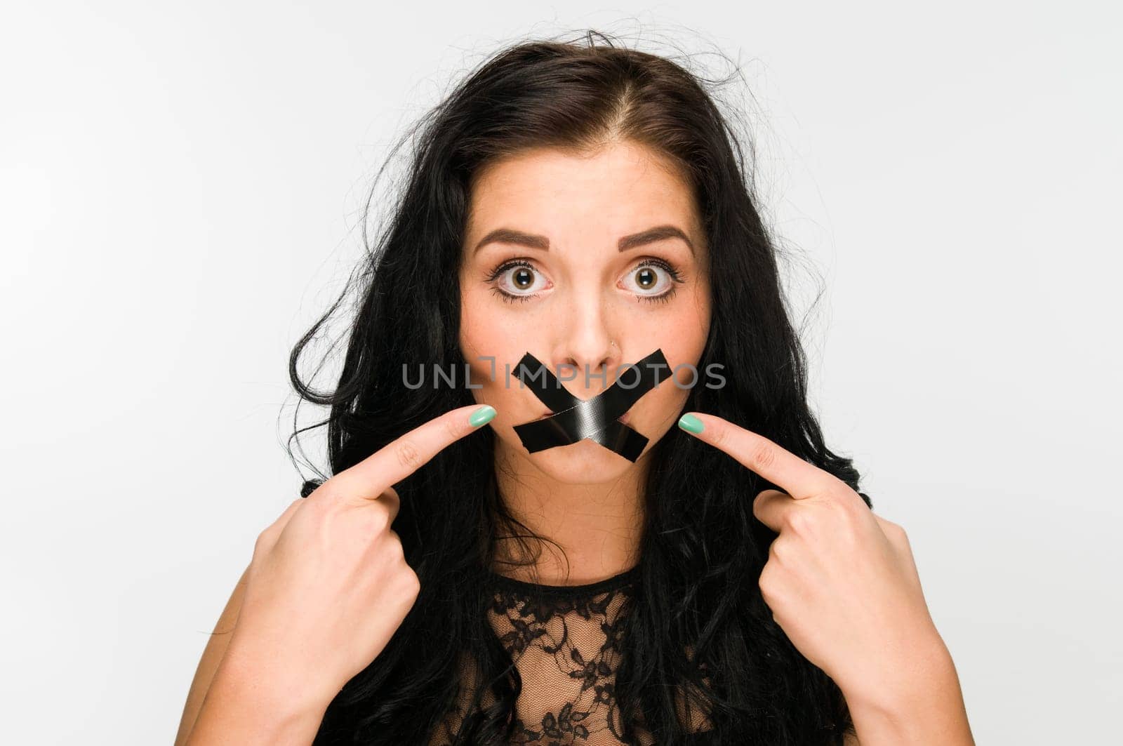 Frontal head-and-shoulders view of a young black-haired woman against a white background with her index fingers pointing to the mouth glued over the cross.