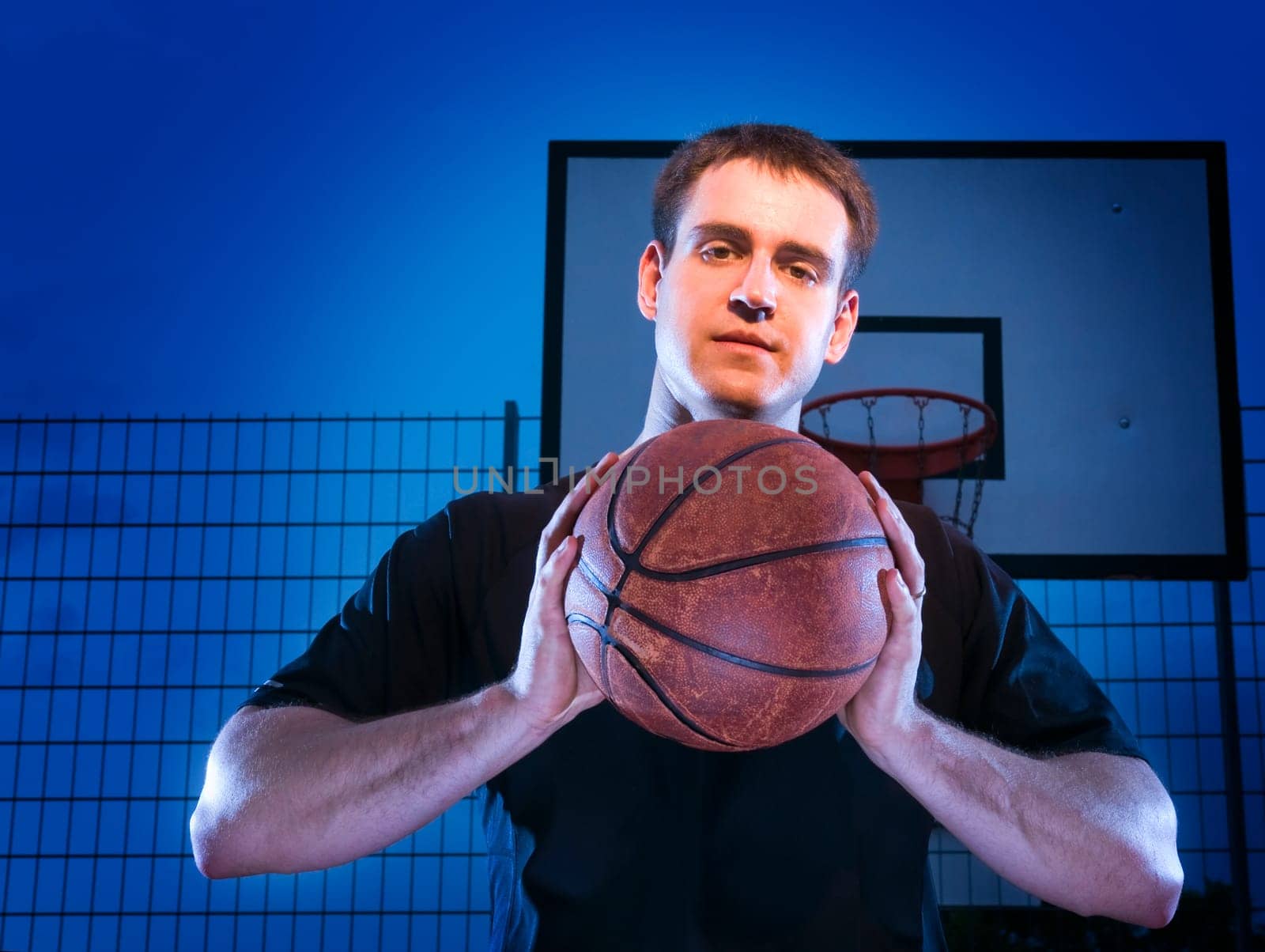 Frontal upper body view of a young man with basketball in front of a blue background at night with basketball hoop.