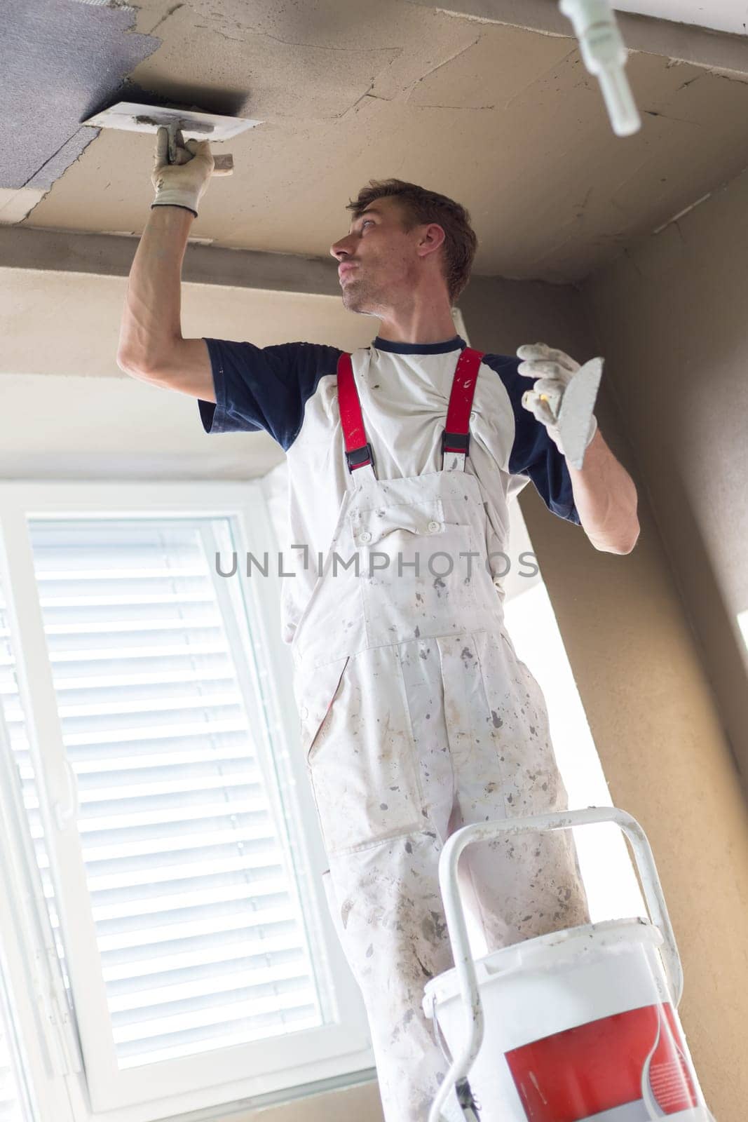 Thirty years old manual worker with wall plastering tools renovating house. Plasterer renovating indoor walls and ceilings with float and plaster.