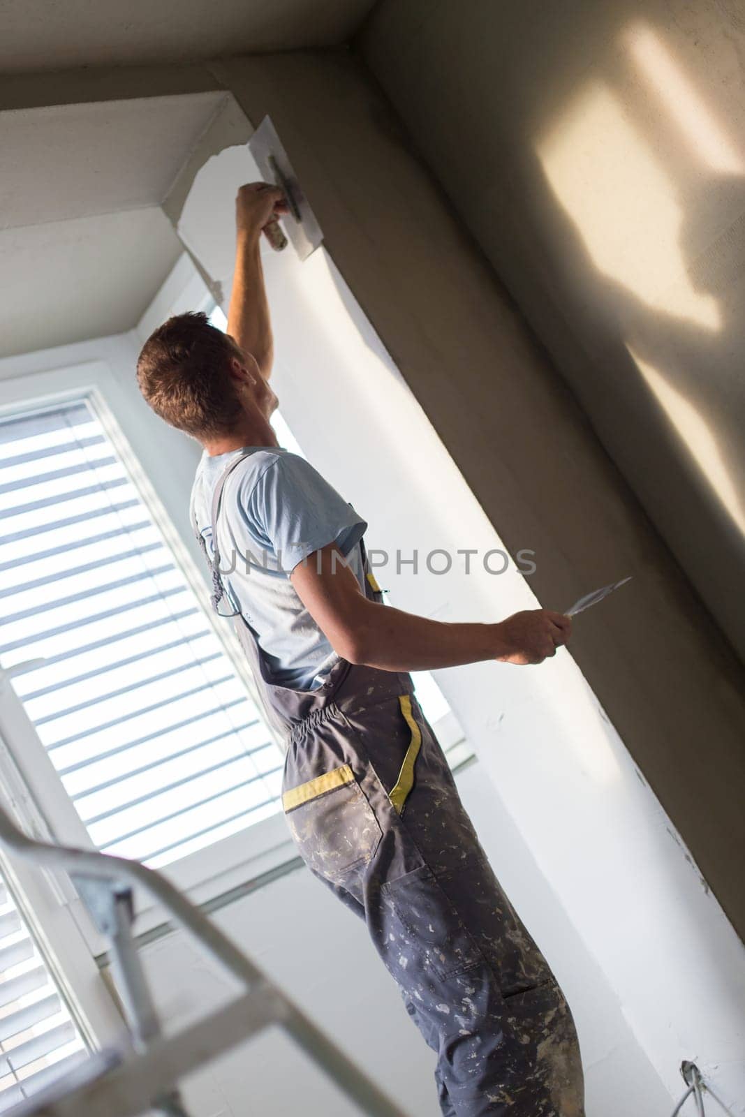 Construction worker wearing worker overall with wall plastering tools renovating apartment house. Plasterer renovating indoor walls and ceilings with float and plaster. Construction finishing works.