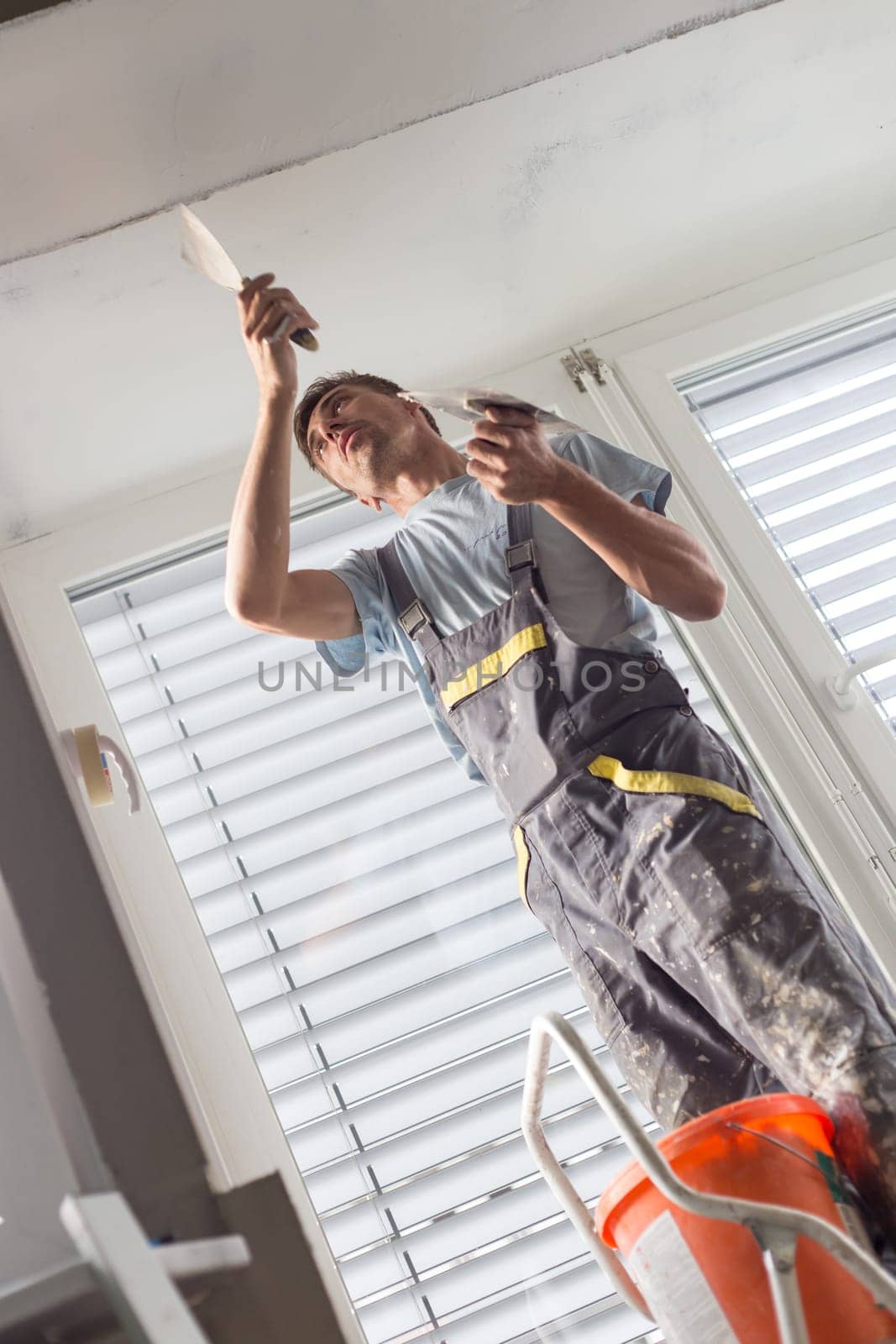 Construction worker wearing worker overall with wall plastering tools renovating apartment house. Plasterer renovating indoor walls and ceilings with float and plaster. Construction finishing works.