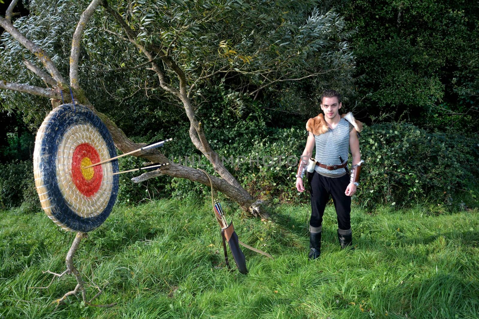Medieval dressed young man stands in nature. Arrow,bow and quiver stands next to it and target made of straw with arrows hangs on the branch on the left side