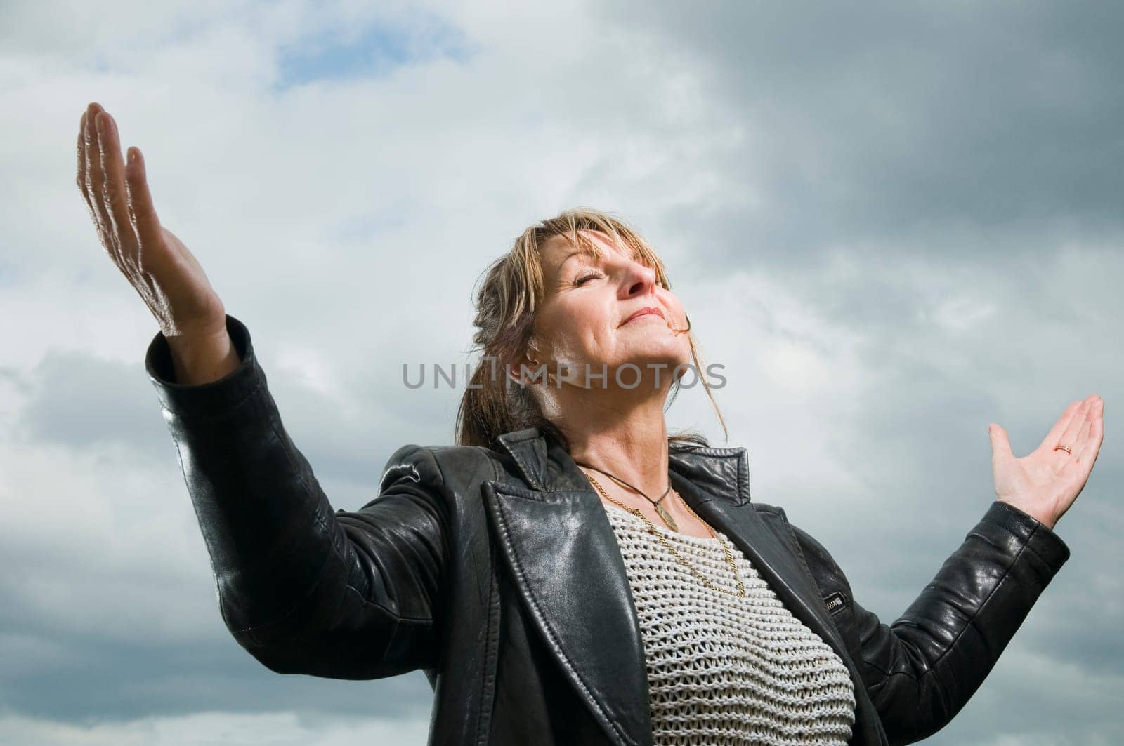 Upper body view of a woman of middle old in front of cloudy sky spreading her arms with her eyes closed.