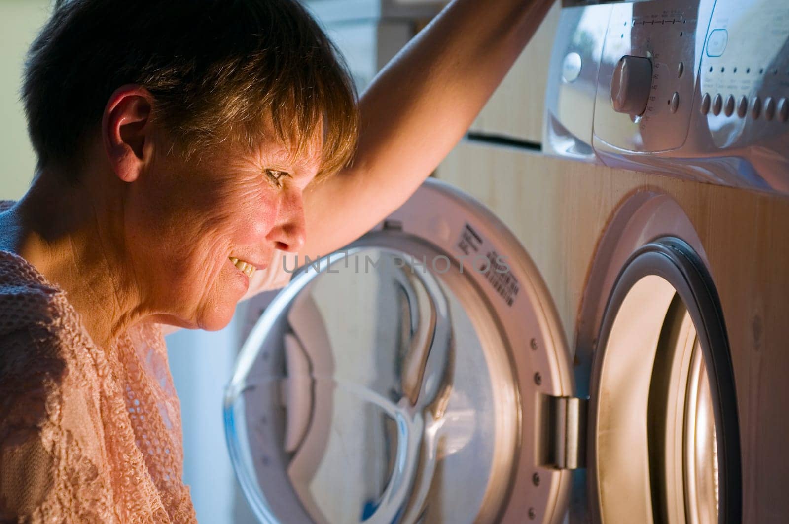 Head-and-shoulder view of a mature woman squatting in front of the open washing machine door and looking into the washing drum.