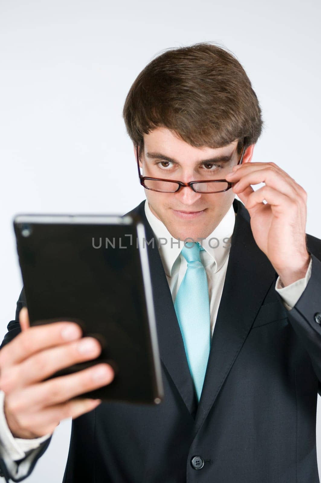 Frontal upper body view of a young man in a dark suit looks over the edge of his glasses at a tablet computer held in his hand against a white background.