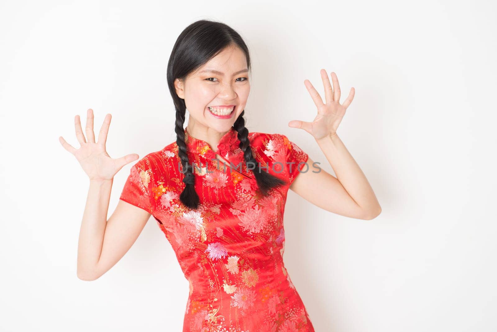 Portrait of young Asian girl in traditional qipao dress with surprised face expression, celebrating Chinese Lunar New Year or spring festival, standing on plain background.