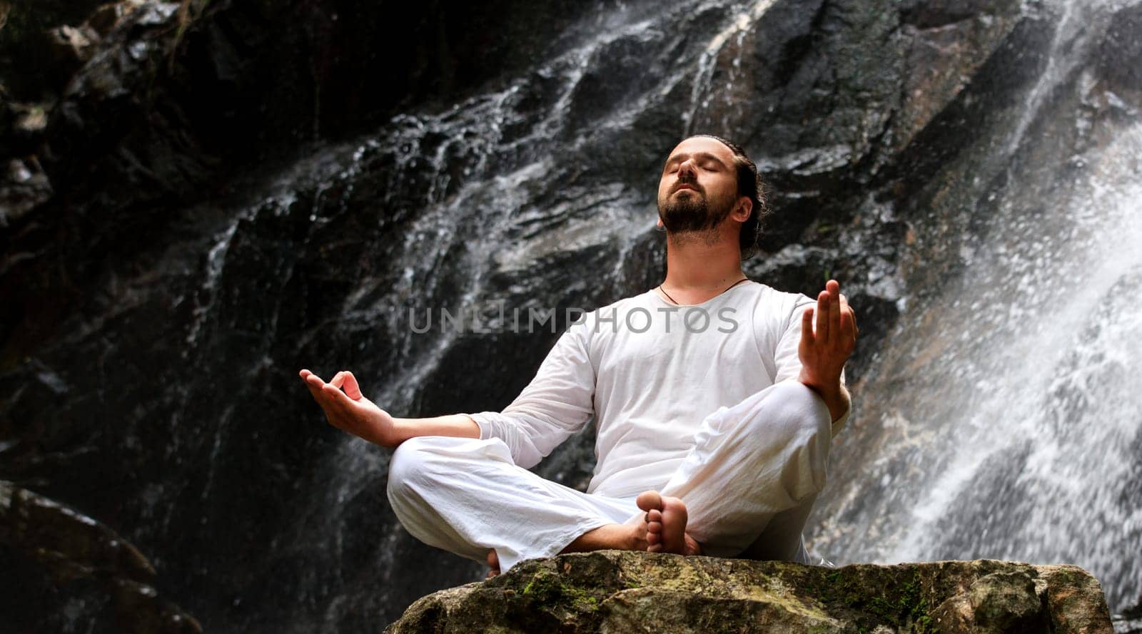 Man sitting in meditation yoga on rock at waterfall in tropical rainforest