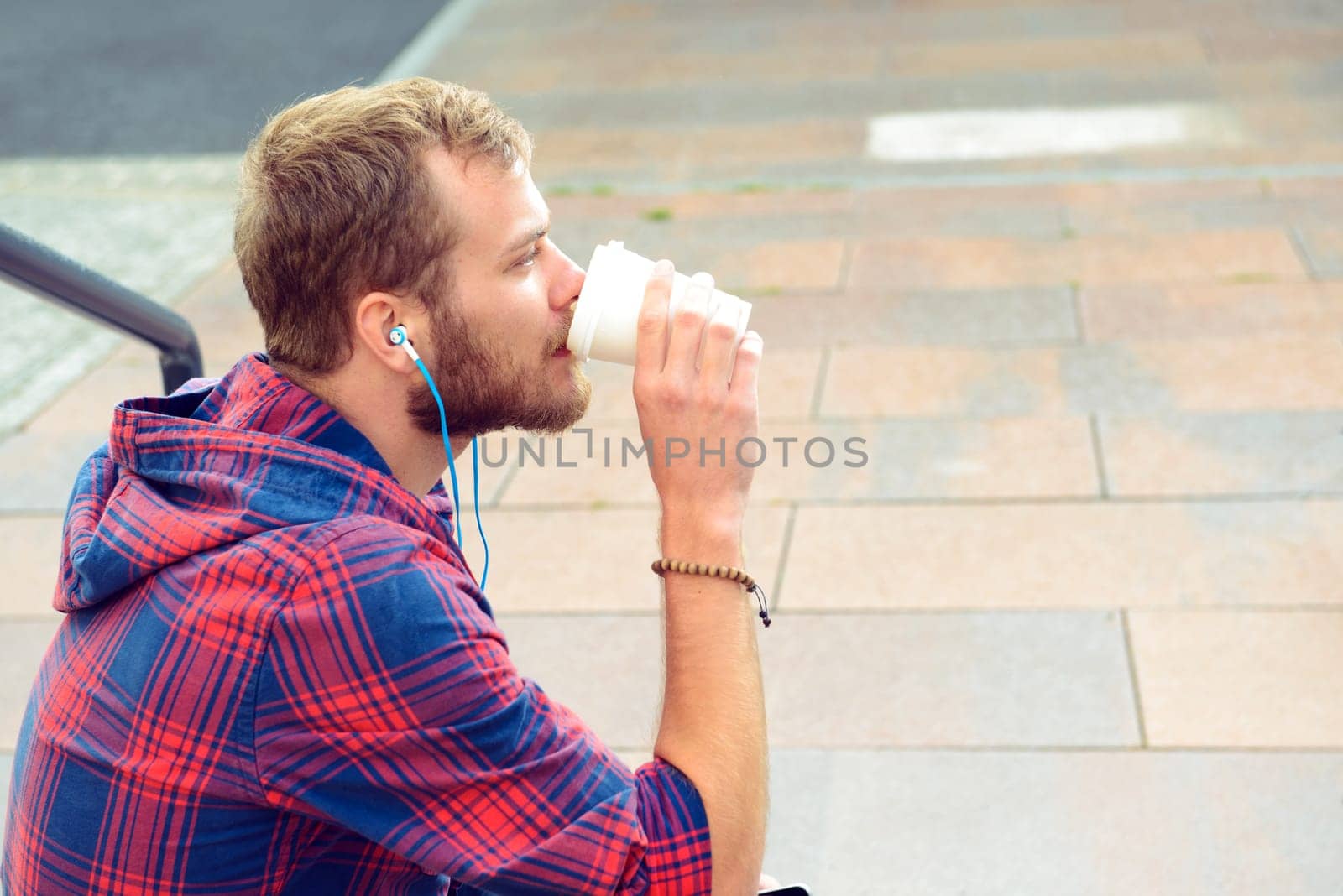 young bearded man in checkered shirt sitting on stairs listening music and drinking soft drink