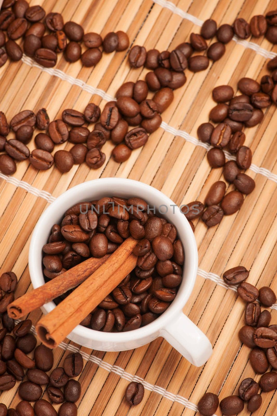 Coffee cup filled with coffee beans and two cinnamon sticks on bamboo mat with ornaments of coffee beans.