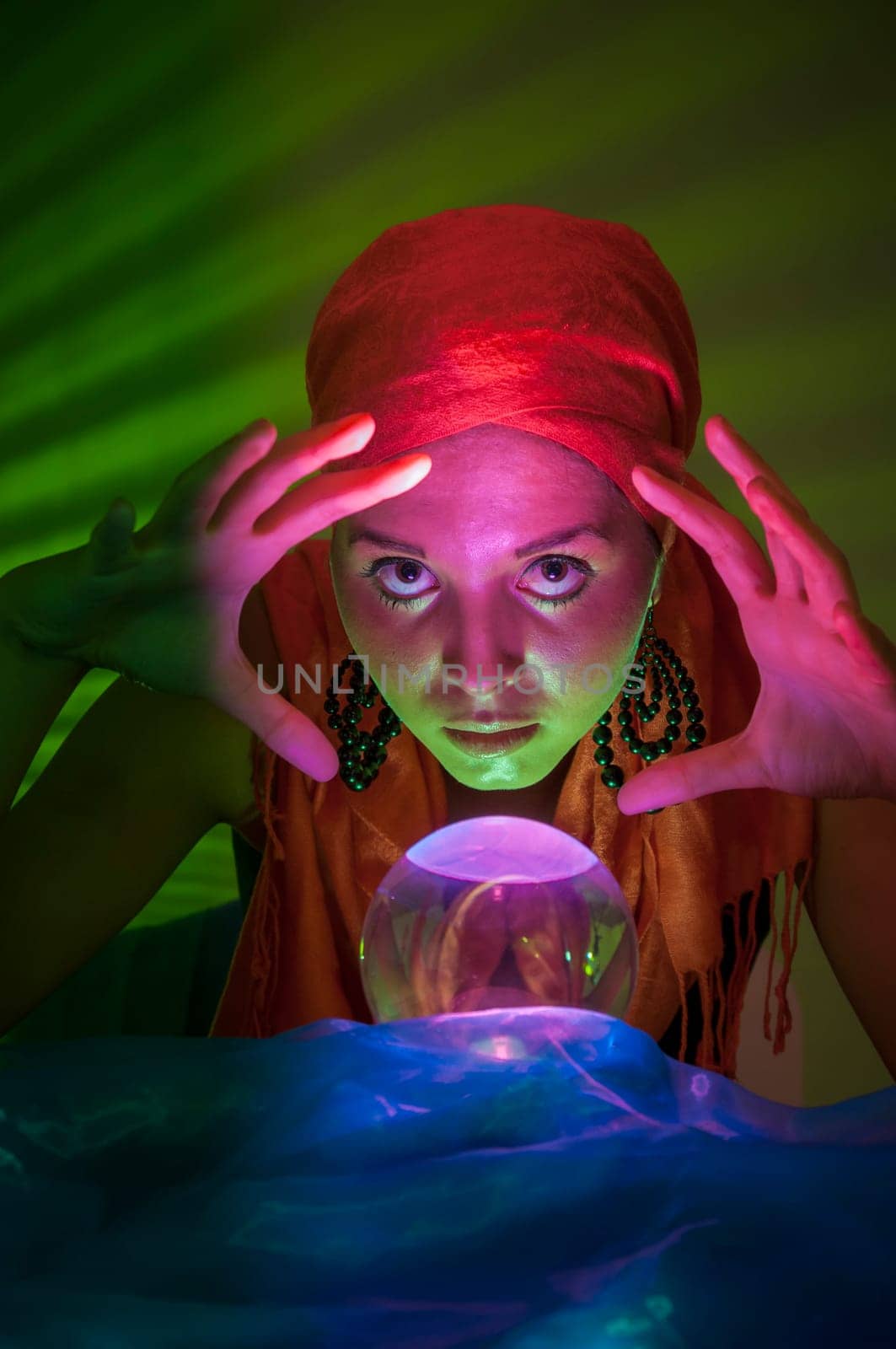 Older female teenager as a fortune teller in gypsy garb in front of a glass ball colorfully illuminated in a dark appearance.