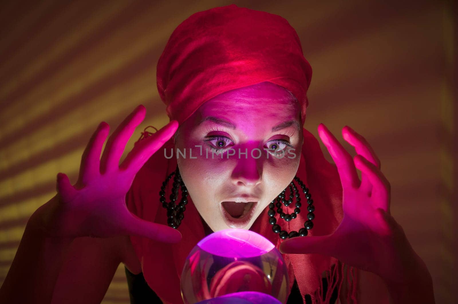 Older female teenager as a fortune teller in gypsy garb in front of a glass ball colorfully illuminated in a dark appearance.