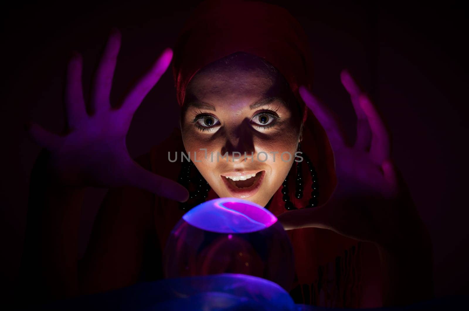 Older female teenager as a fortune teller in gypsy garb in front of a glass ball colorfully illuminated in a dark appearance.