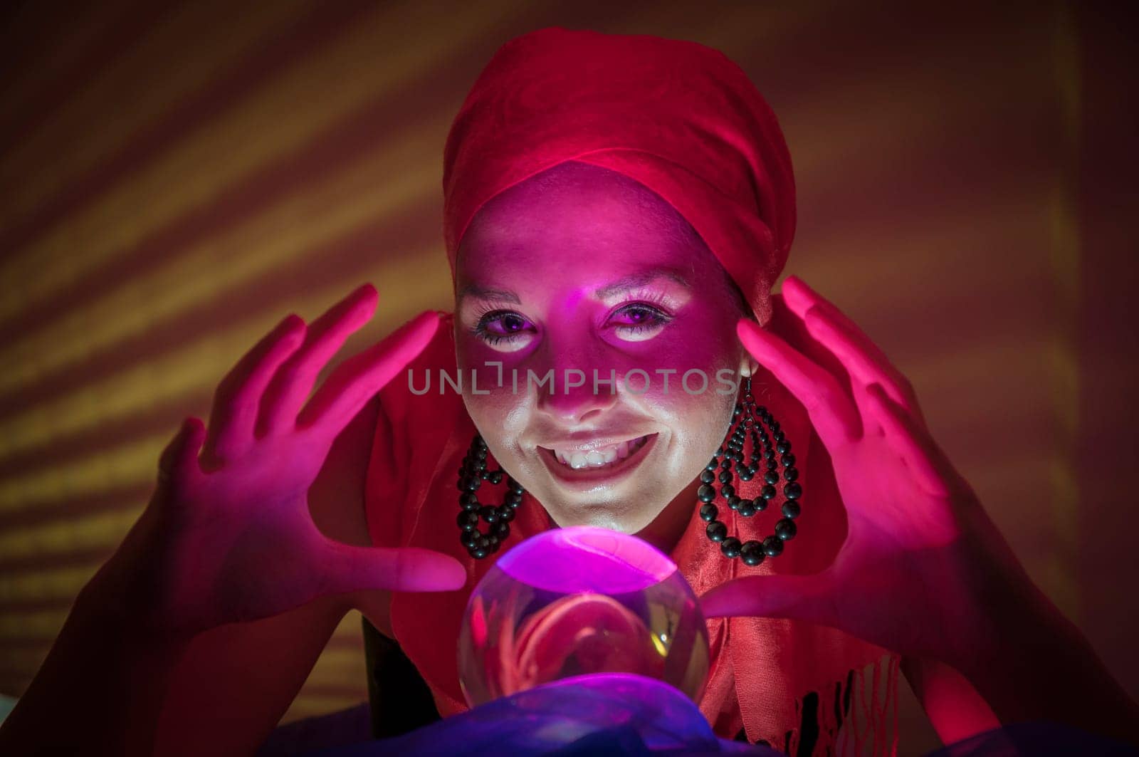 Older female teenager as a fortune teller in gypsy garb in front of a glass ball colorfully illuminated in a dark appearance.