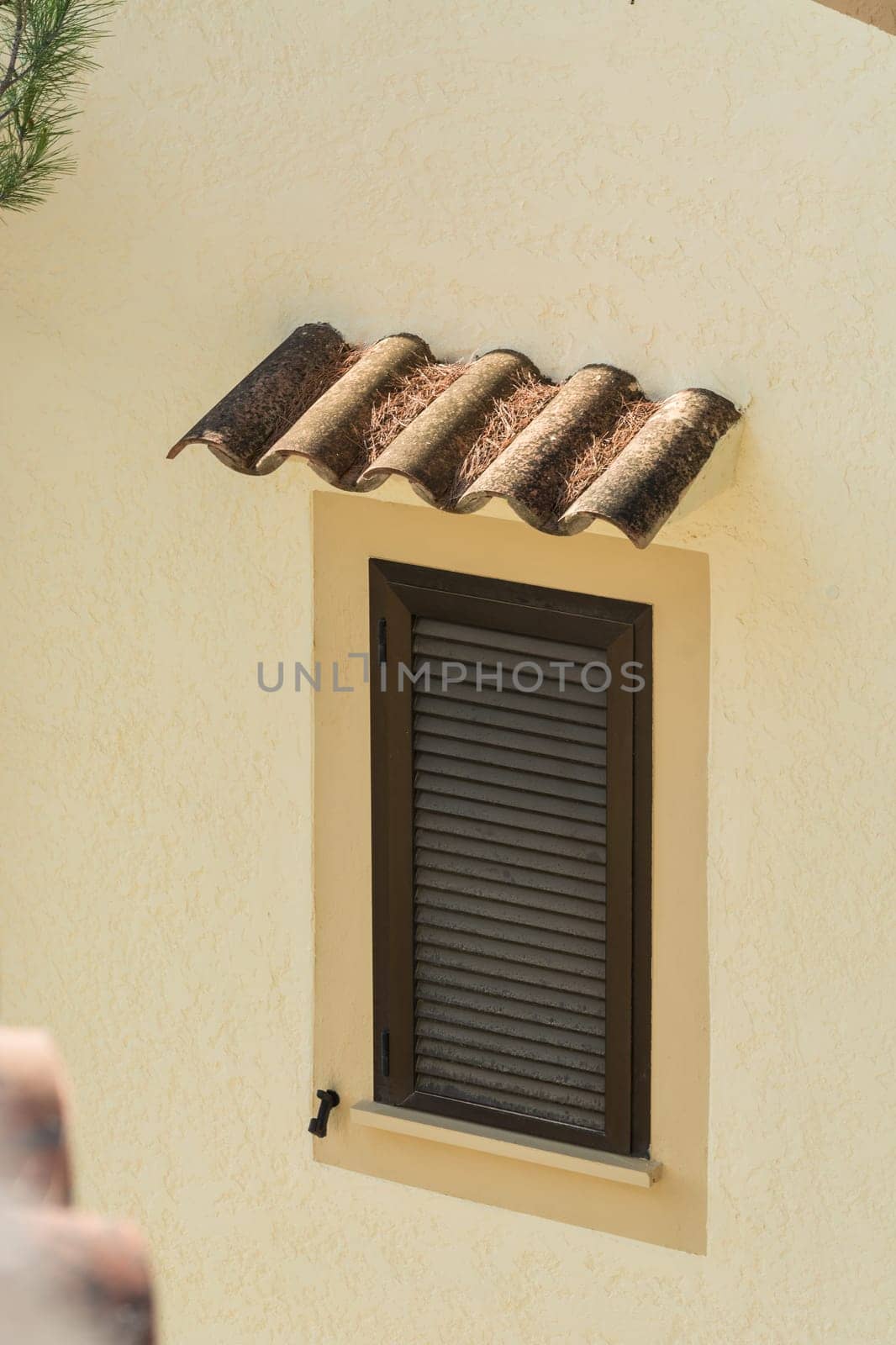 Window with brown closed shutter. Rain cover made of roof tiles in a yellow painted house wall..
