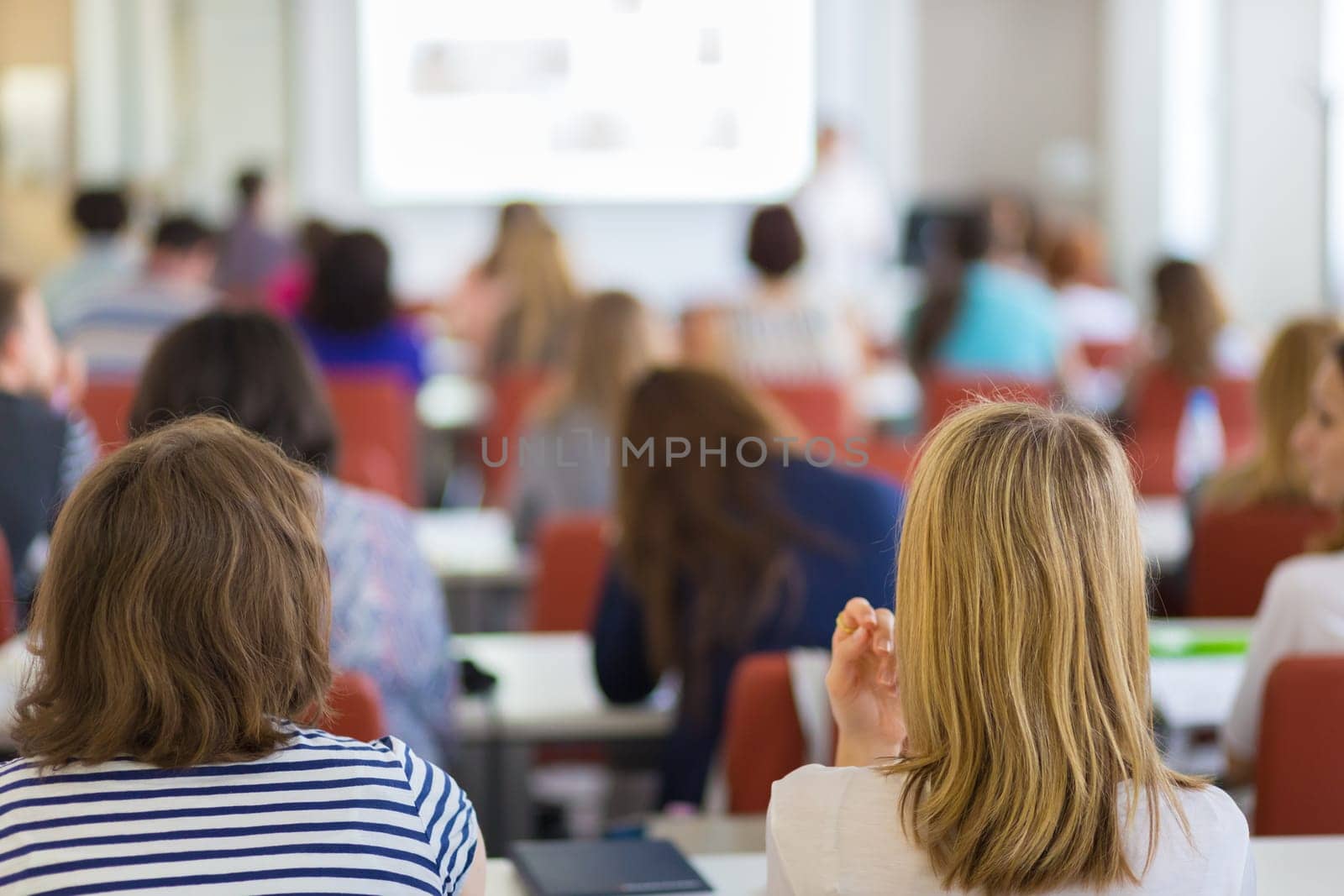 Speaker giving presentation in lecture hall at university. Participants listening to lecture and making notes.
