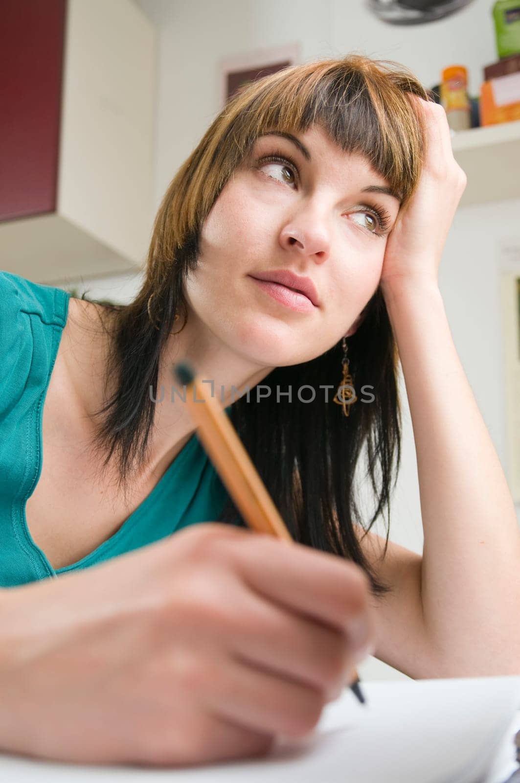 Wide-angle upper body view of a young brunette woman sitting at the home table with pen in hand looking to the side.